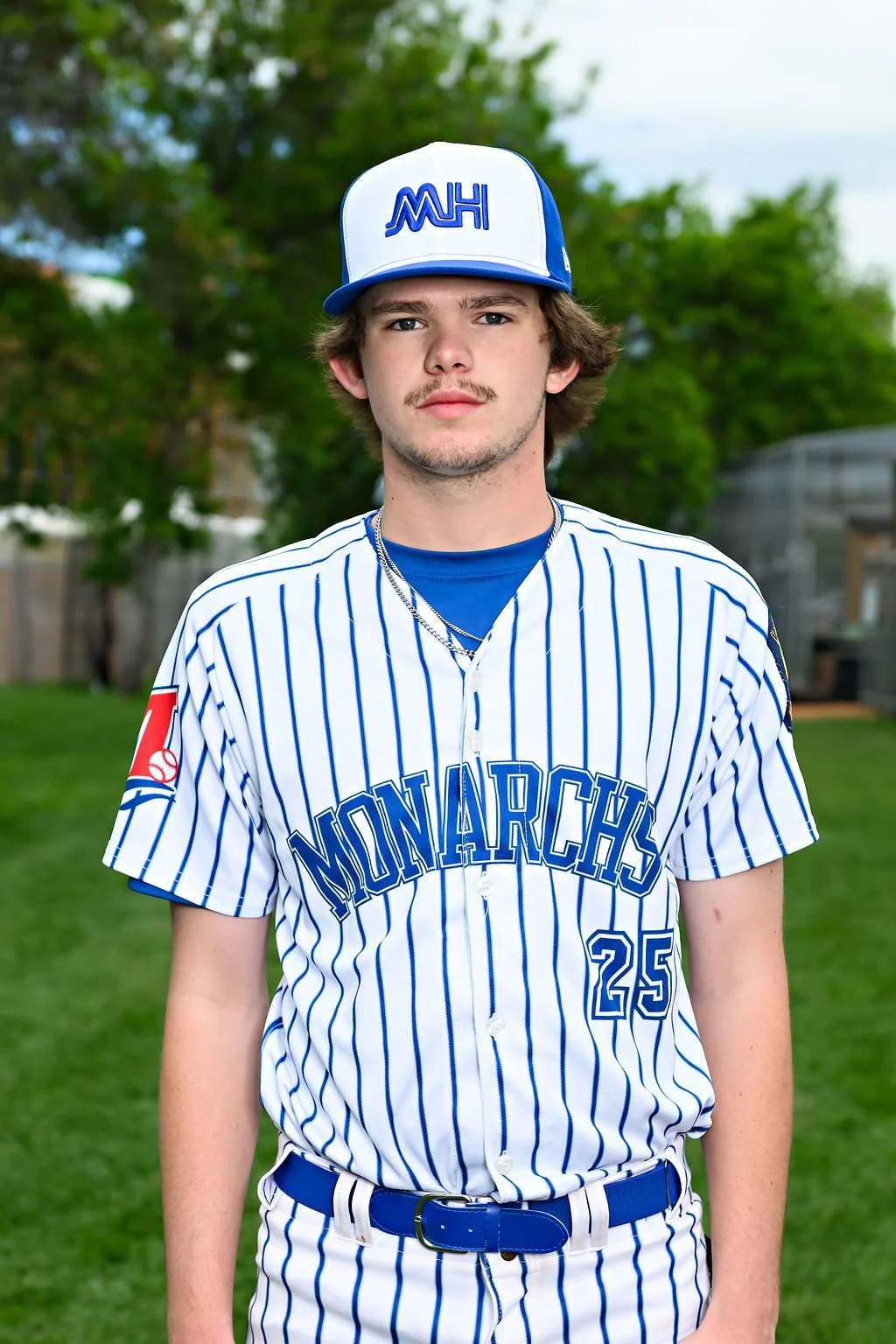 Young male baseball player wearing a blue jersey and a white cap with blue lettering, standing on a baseball field.