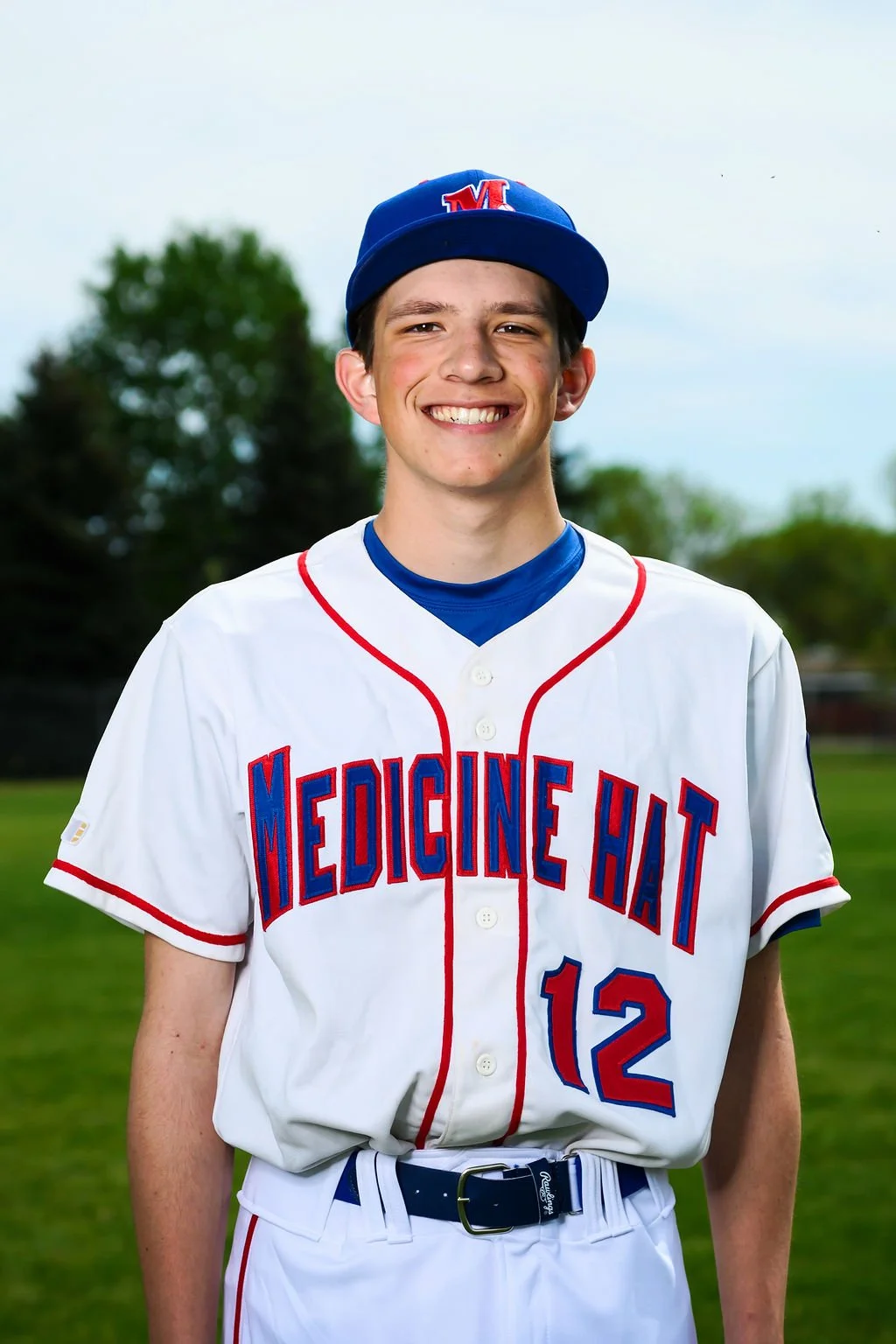 A young male baseball player in a white uniform with red and blue accents, wearing a blue cap with a red and white 'M' logo, holding a black and tan baseball glove, standing outdoors on a baseball field with trees and an overcast sky in the background.
