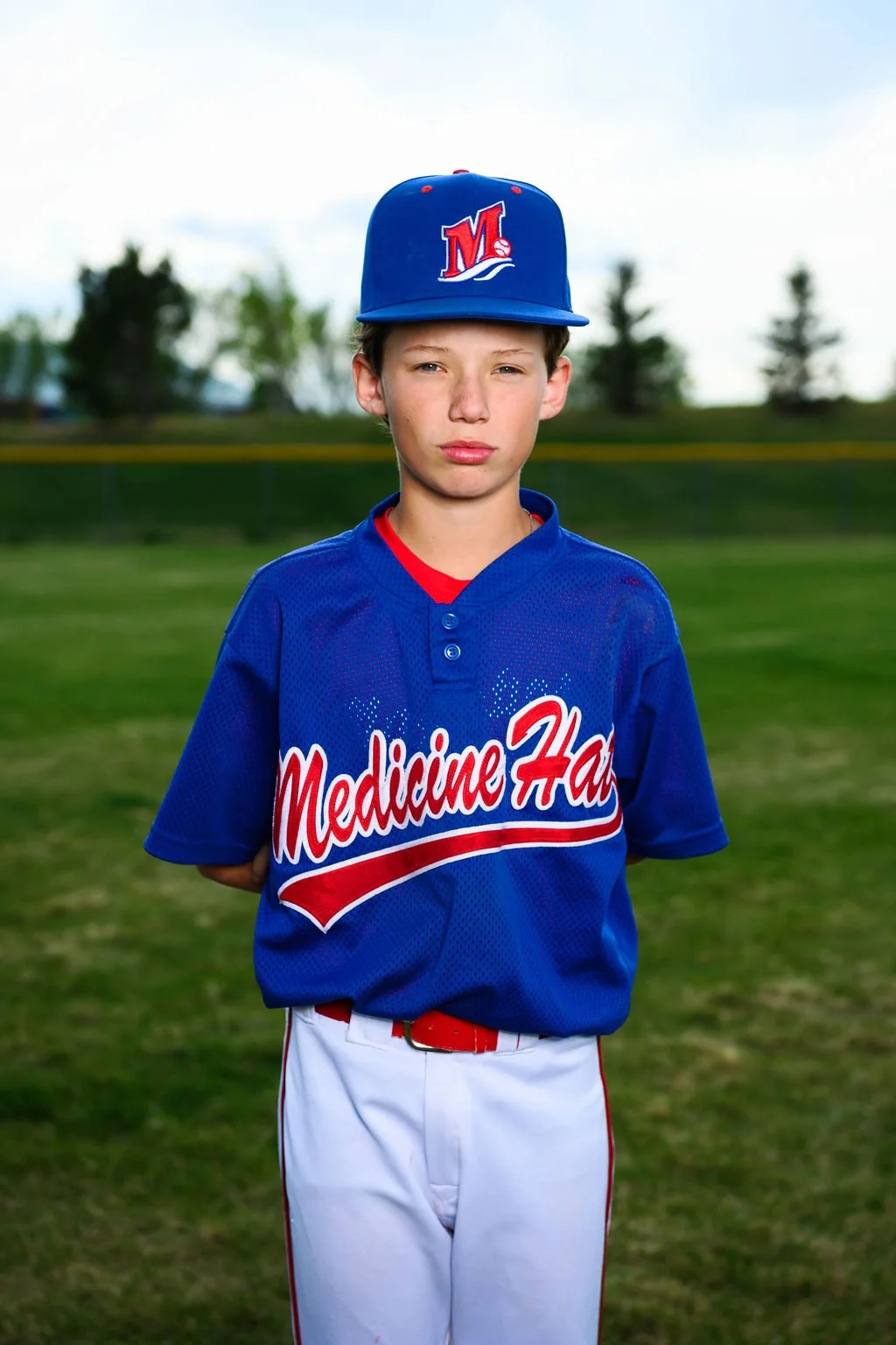 A young boy wearing a blue baseball jersey with 'Medicine Hat' written on it and a matching blue cap with a logo, standing outdoors on a field with trees in the background.