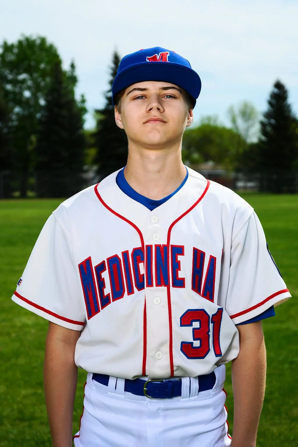 A young male baseball player in a white uniform with red and blue lettering that reads 'Medicine H', wearing a blue baseball cap with a red 'M' logo and holding a baseball glove on a baseball field.