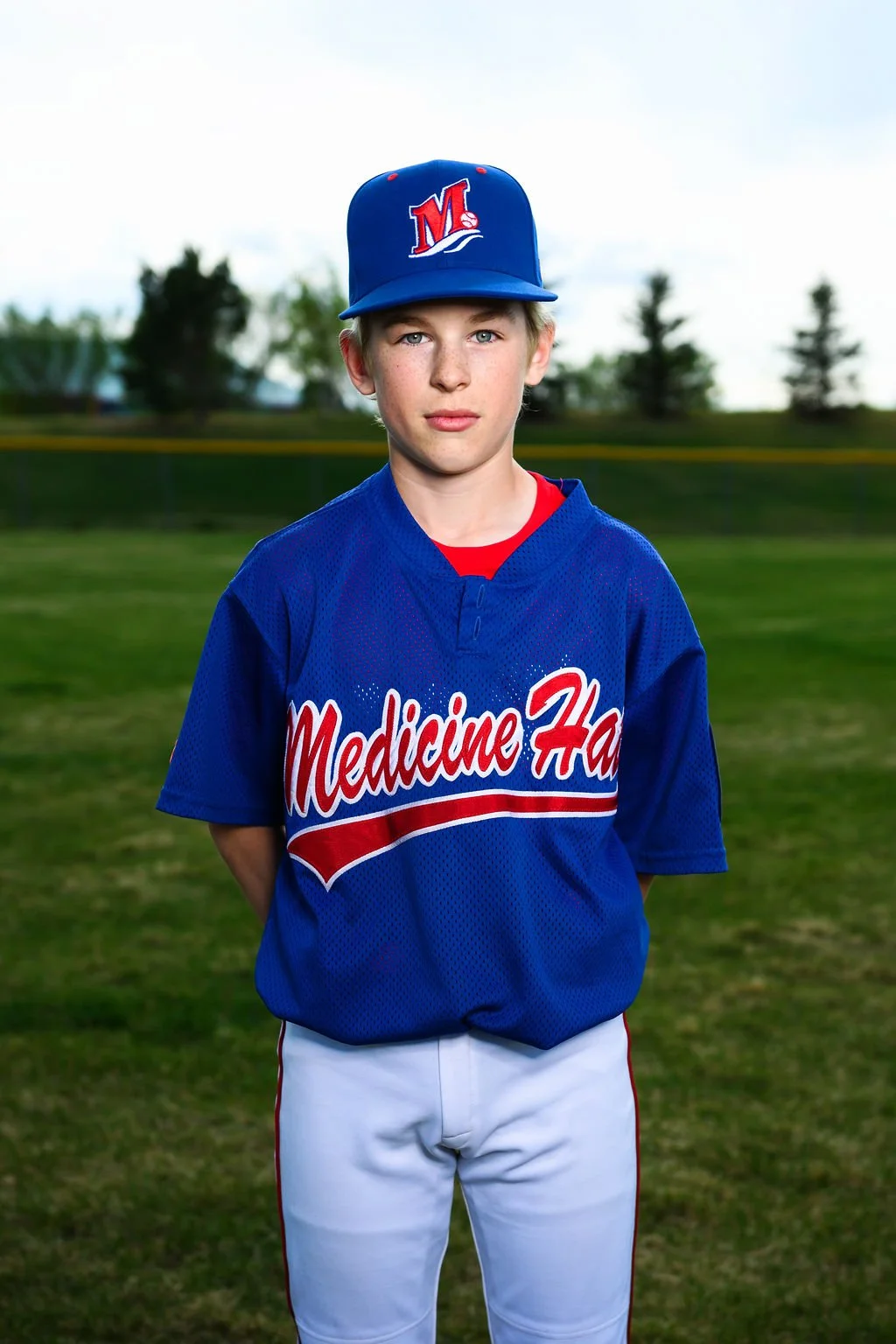 A young man wearing a blue baseball uniform and cap, standing outdoors with trees and a clear sky in the background.