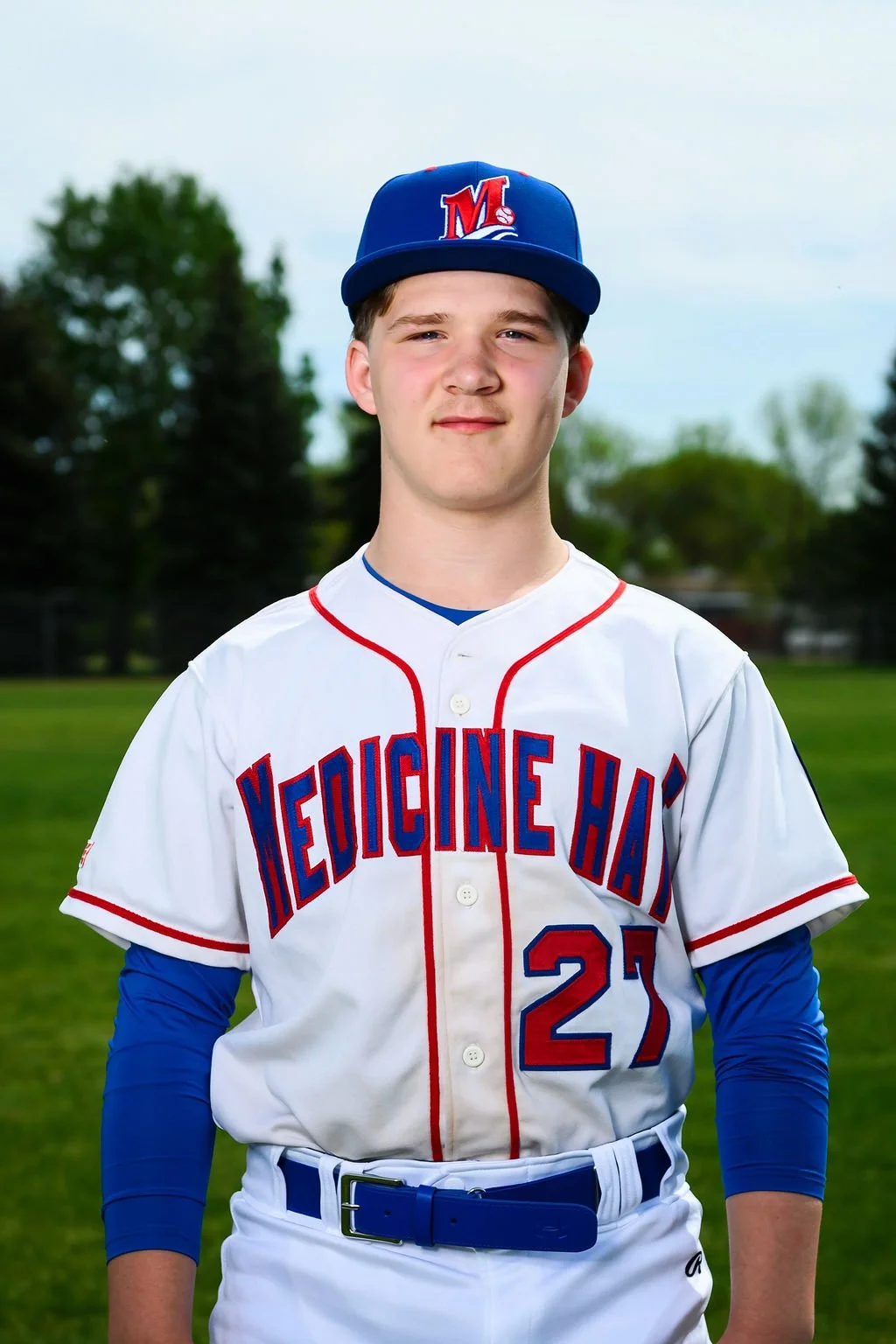 Young baseball player in a blue cap and white jersey with red and blue lettering, holding a glove on a baseball field during daytime.