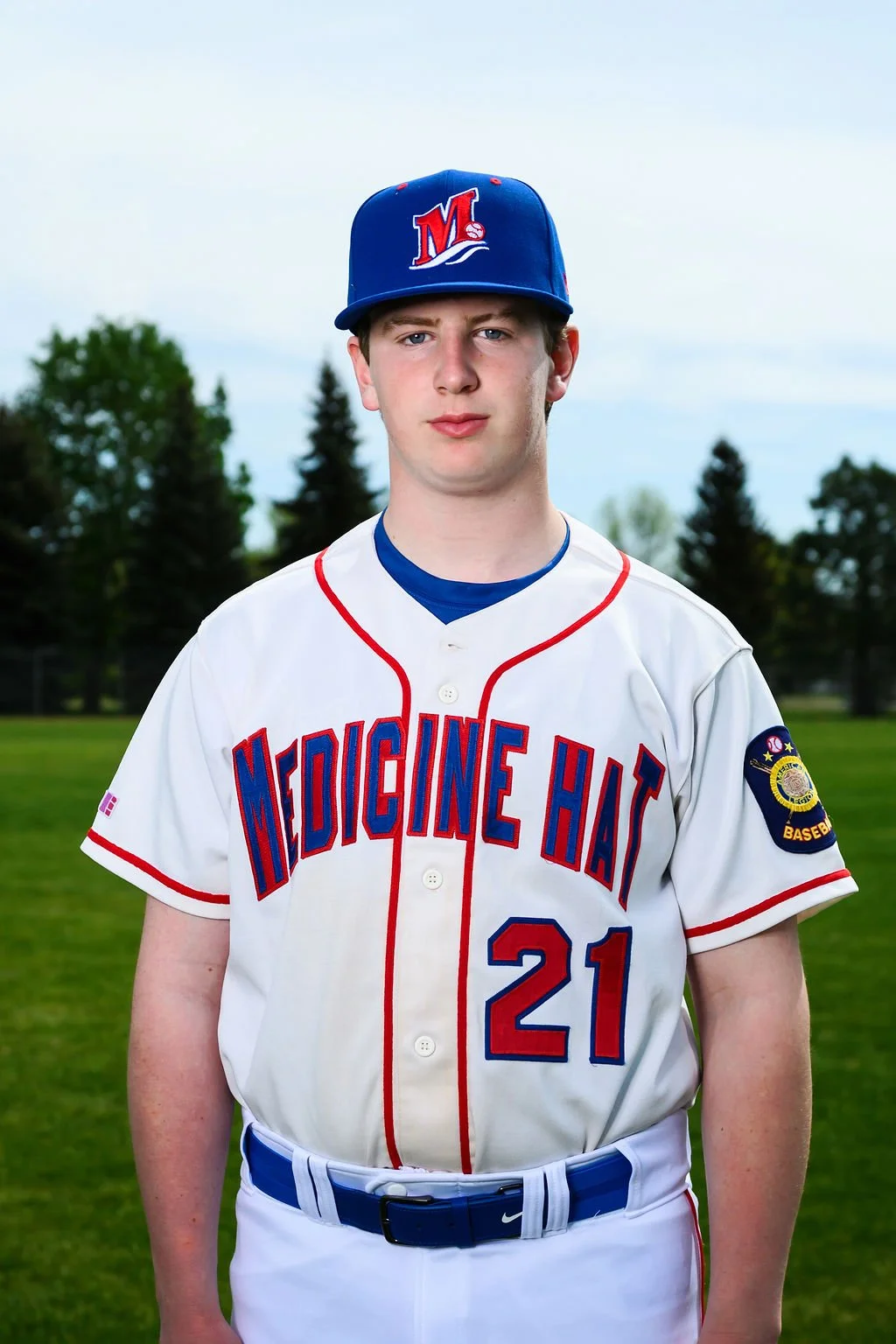 Young baseball player in a white jersey with red and blue accents, a blue cap with a red and white logo, holding a baseball glove, standing on a baseball field with trees and a cloudy sky in the background.