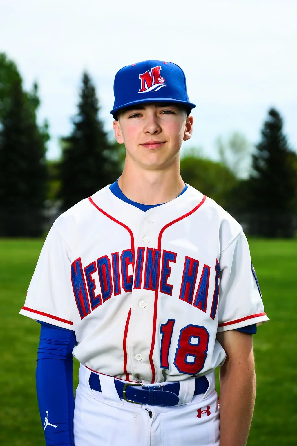 A teenage baseball player in a Medcine team uniform standing on the field, holding a baseball glove, with trees and cloudy sky in the background.