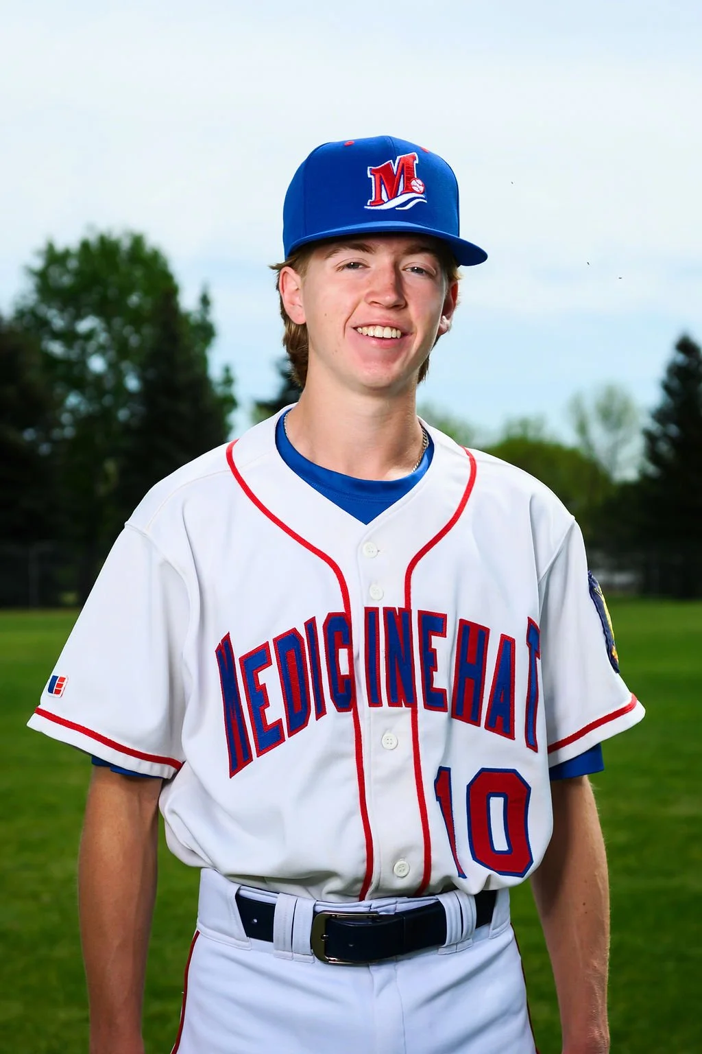 Young baseball player in a white uniform with red and blue lettering, wearing a blue cap, standing on a baseball field with trees in the background.