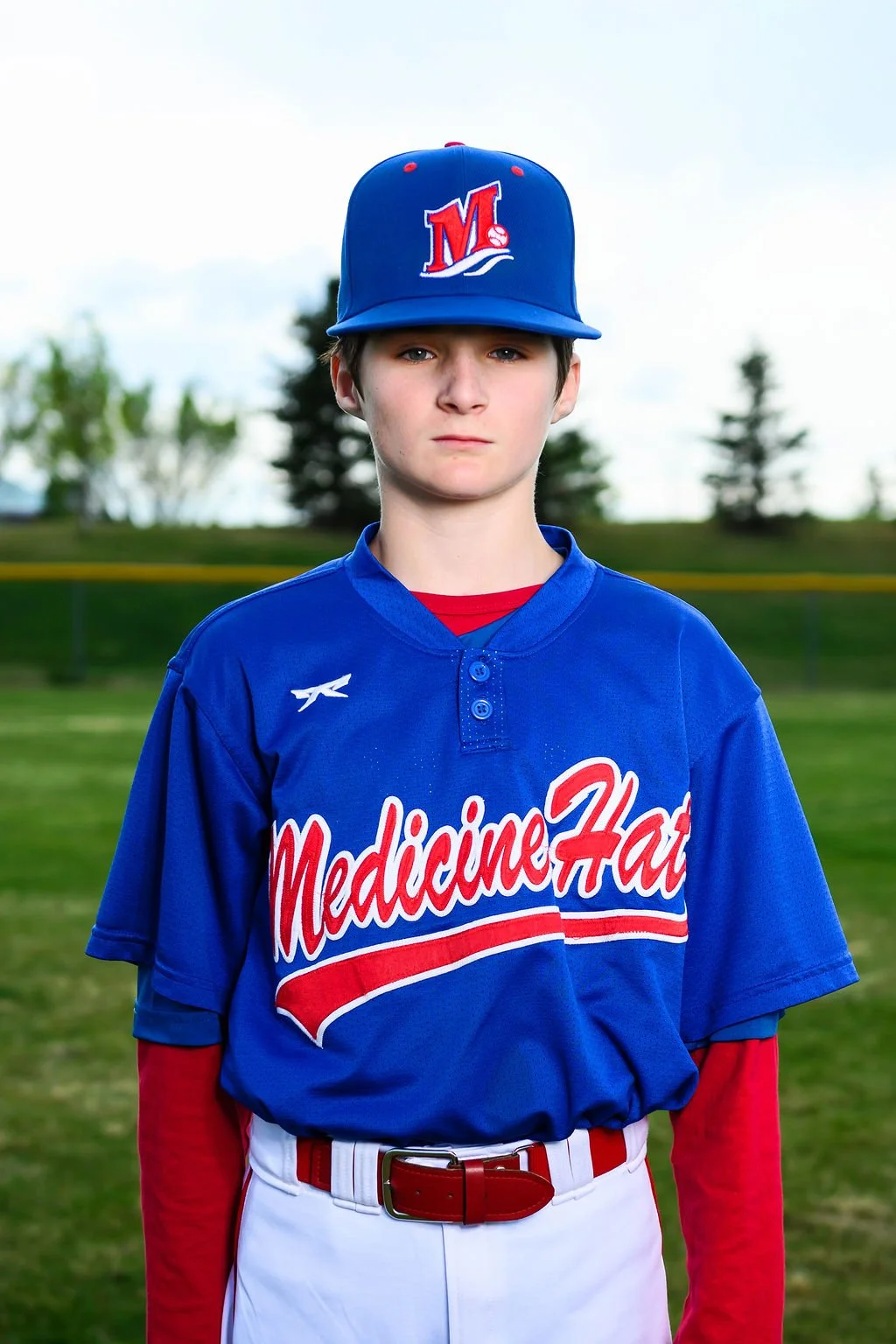 A teenage baseball player with blond hair and blue eyes wearing a blue jersey with 'Medicine Hat' written on it, and a matching blue cap with a red and white logo, standing outdoors on a grassy field with pine trees in the background.