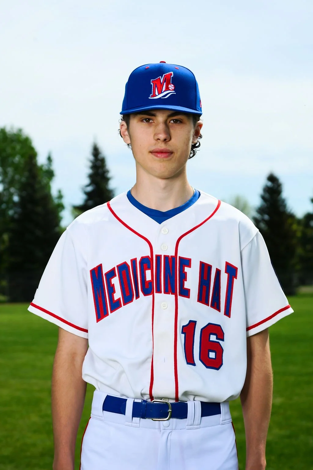 A young male baseball player in uniform standing on a baseball field, holding a baseball glove, with an overcast sky in the background.