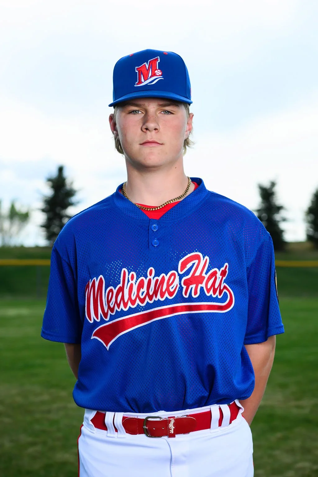 A young baseball player wearing a blue jersey with 'Medicine Hat' written on it, a blue cap with a red and white 'M' logo, and sports sunglasses, standing outdoors with a background of trees and a blue sky.