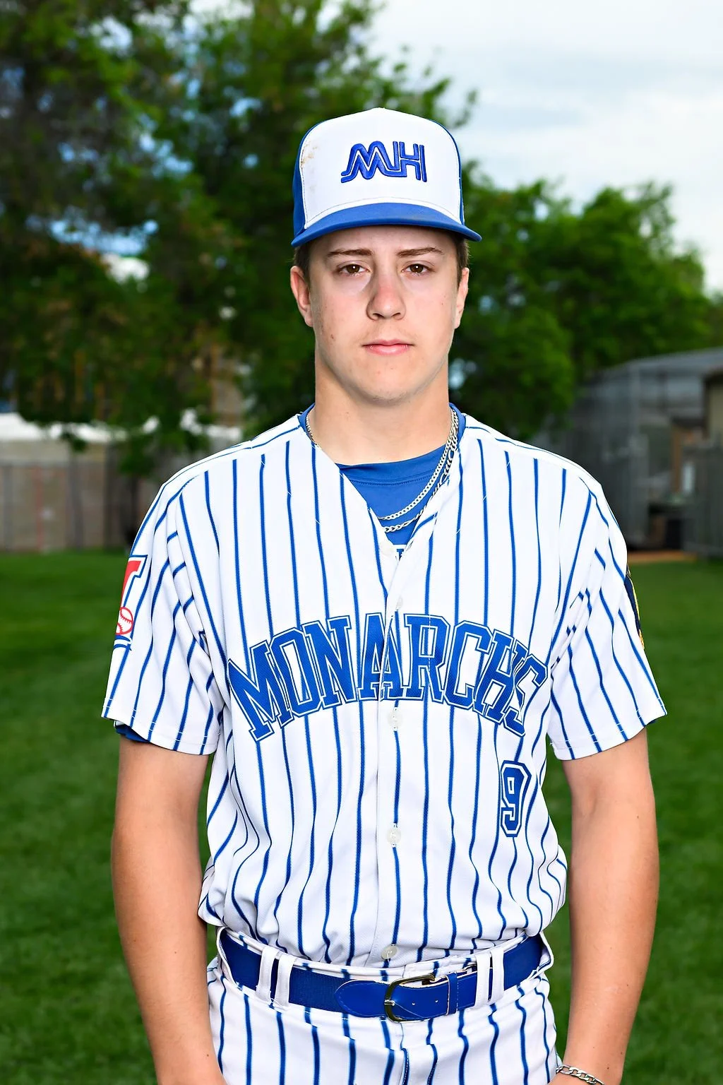 Young man in blue baseball uniform standing on a field, wearing a white cap with blue writing, on an outdoor baseball field with trees and a clear sky in the background.