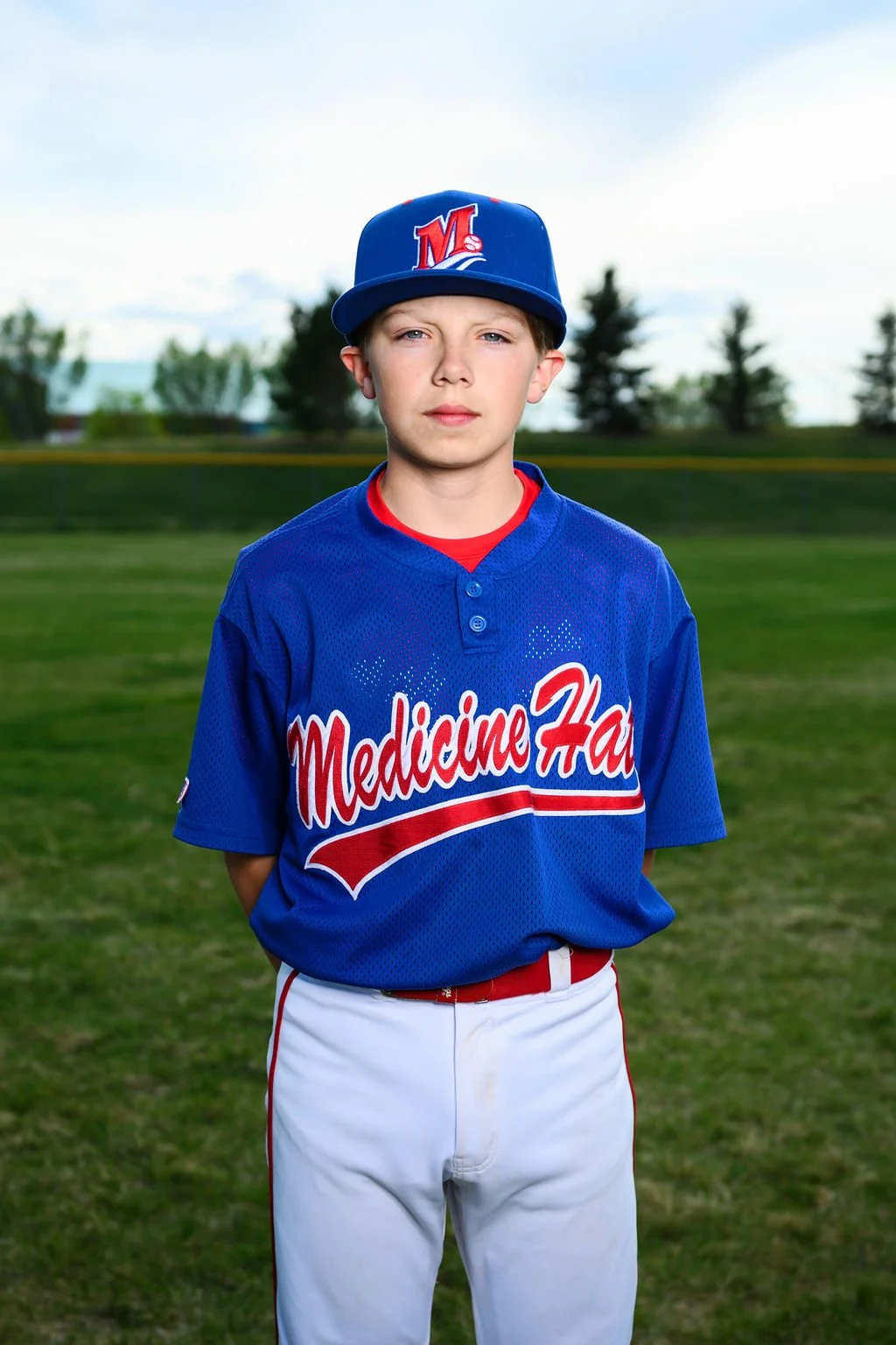 Young male baseball player wearing a blue jersey with 'Medicine Hat' written across it and a matching blue cap with red and white embroidery, standing outdoors with trees and a partly cloudy sky in the background.