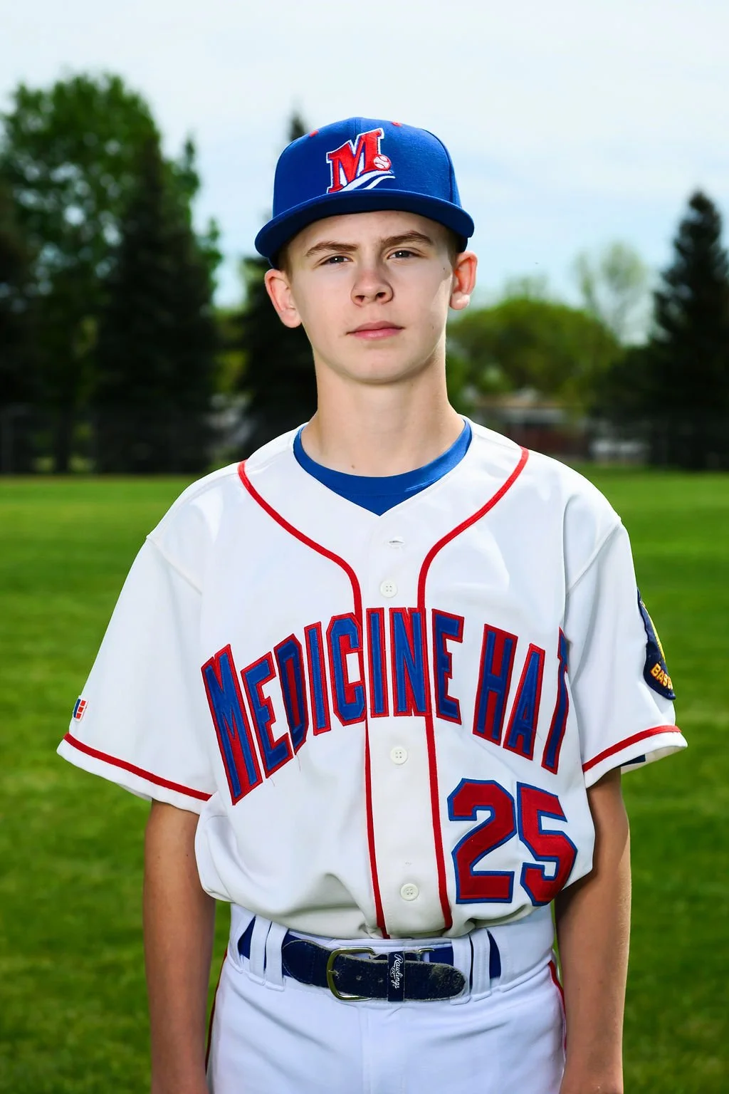 A young baseball player standing on a field with a baseball glove, wearing a white uniform with red and blue accents, a blue cap with a red and white logo, and a necklace, with trees and a cloudy sky in the background.