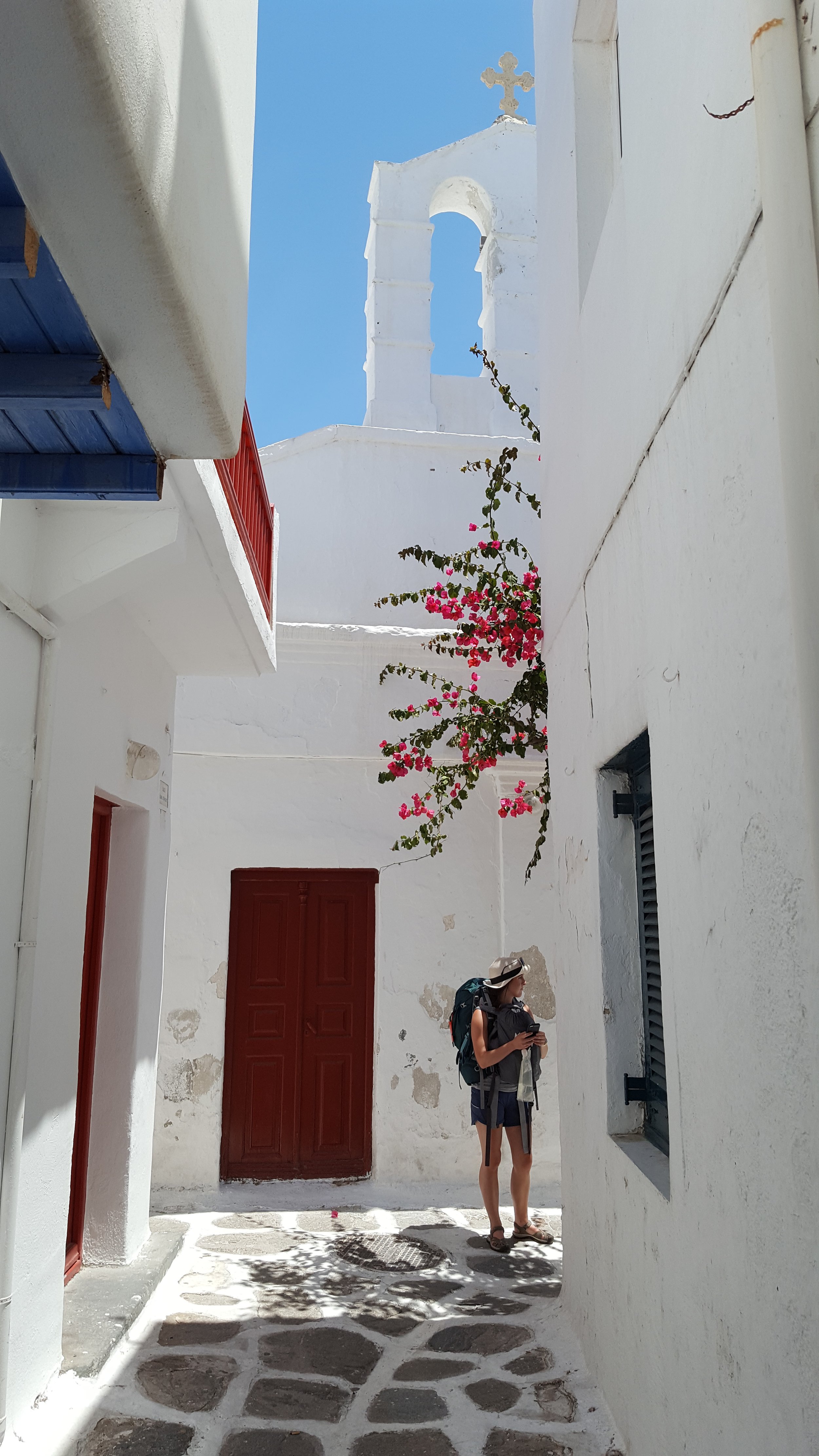 Anne stands on a corner in Mykonos, Greece. She is wearing a straw hat, shorts, and a backpack, looking down the lane.