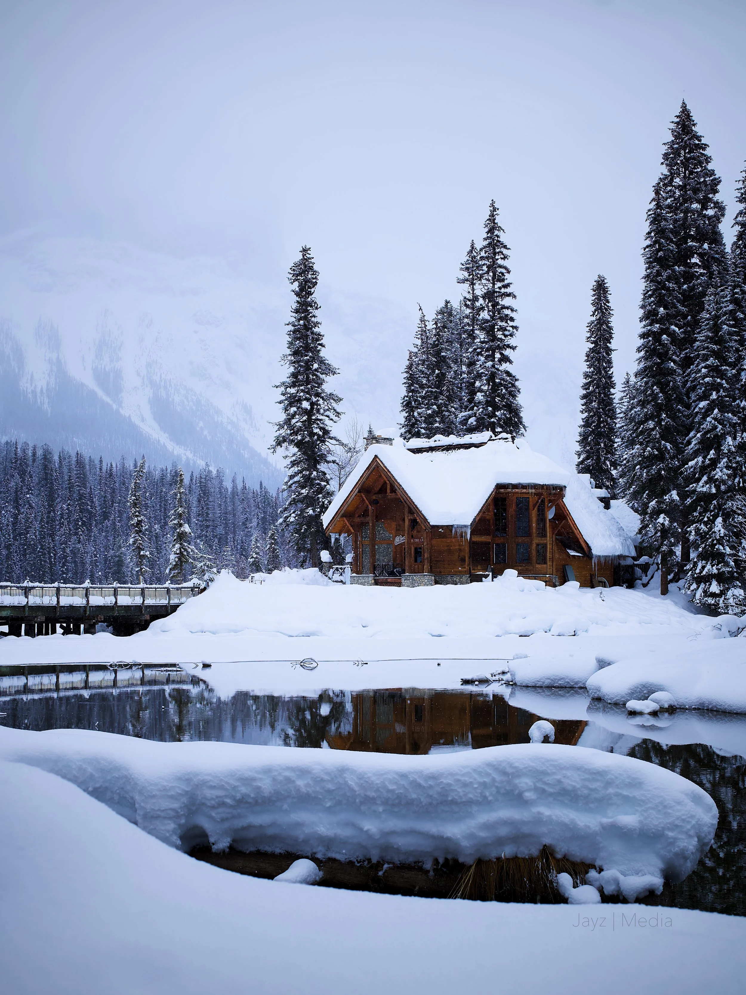 A wooden cabin surrounded by snow-covered trees and landscape, with a river in the foreground reflecting the scene, in a winter mountain setting.