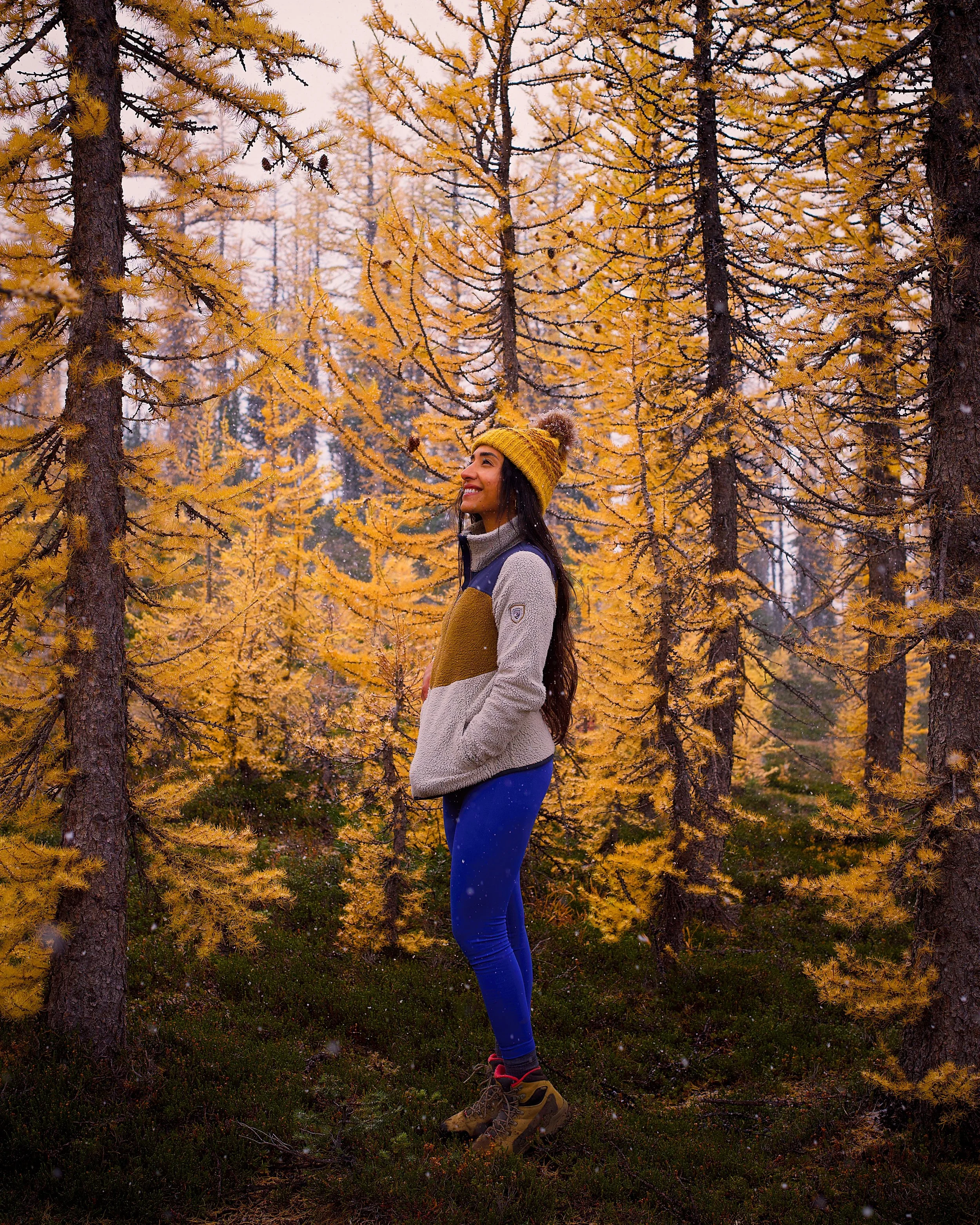 A woman standing in a forest of yellow autumn trees, smiling and looking to the side, wearing a yellow knit hat, a multicolored fleece jacket, blue leggings, and hiking boots.