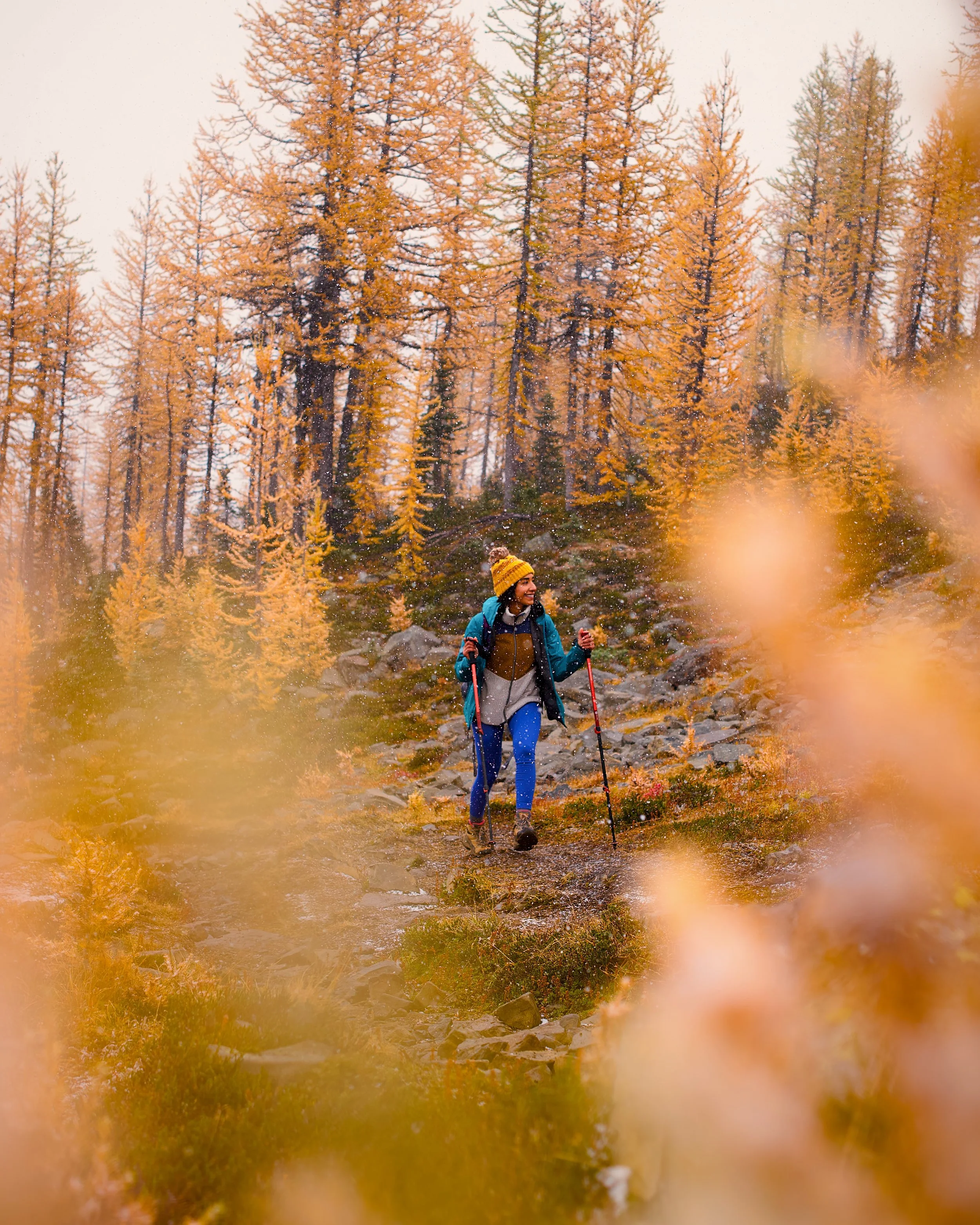 A woman hiking outdoors in autumn, wearing a yellow beanie, teal jacket, and blue leggings, using trekking poles among orange and yellow trees.