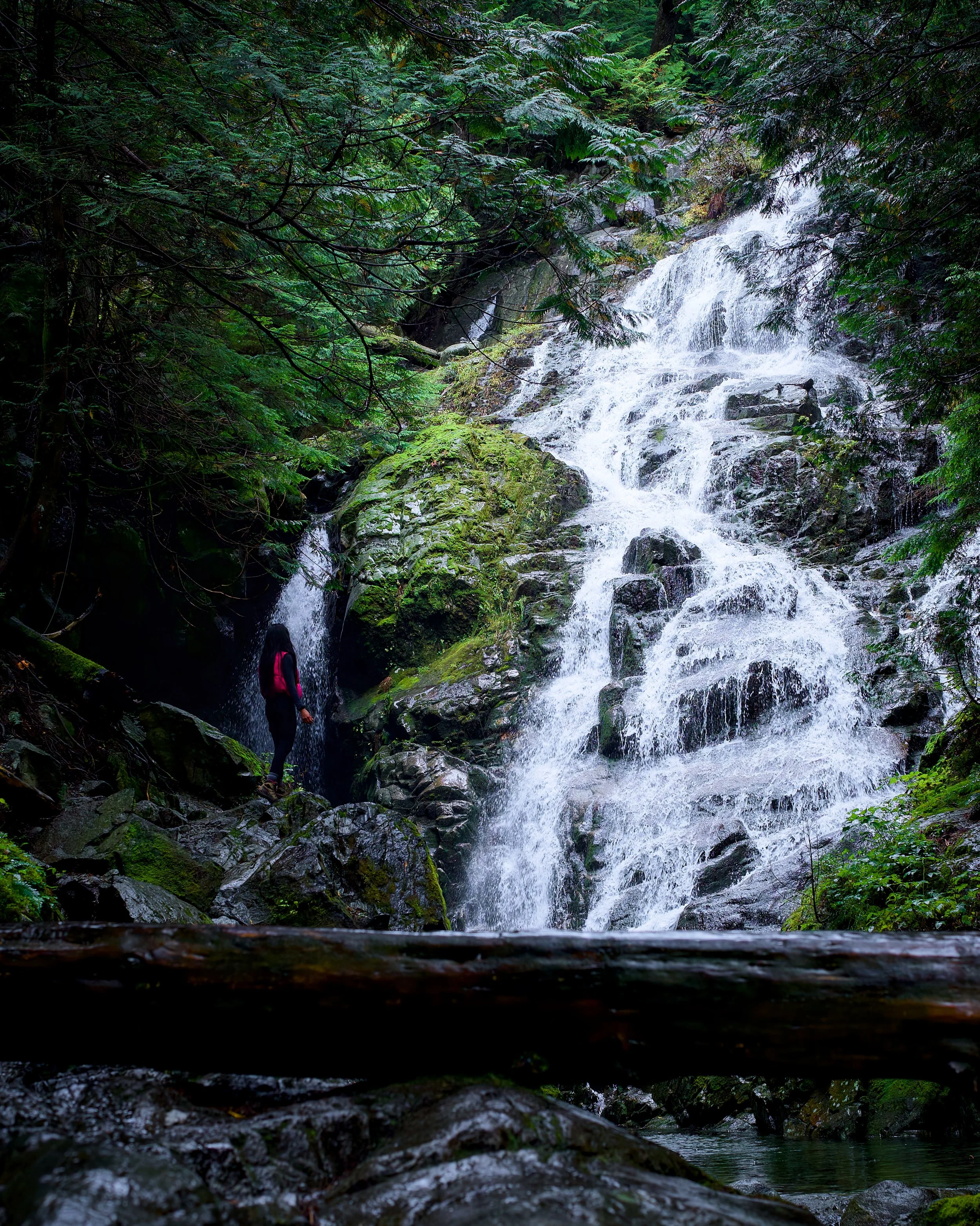 A hiker in dark clothing and a red backpack stands near the base of a cascading waterfall in a lush green forest, surrounded by moss-covered rocks and dense foliage.