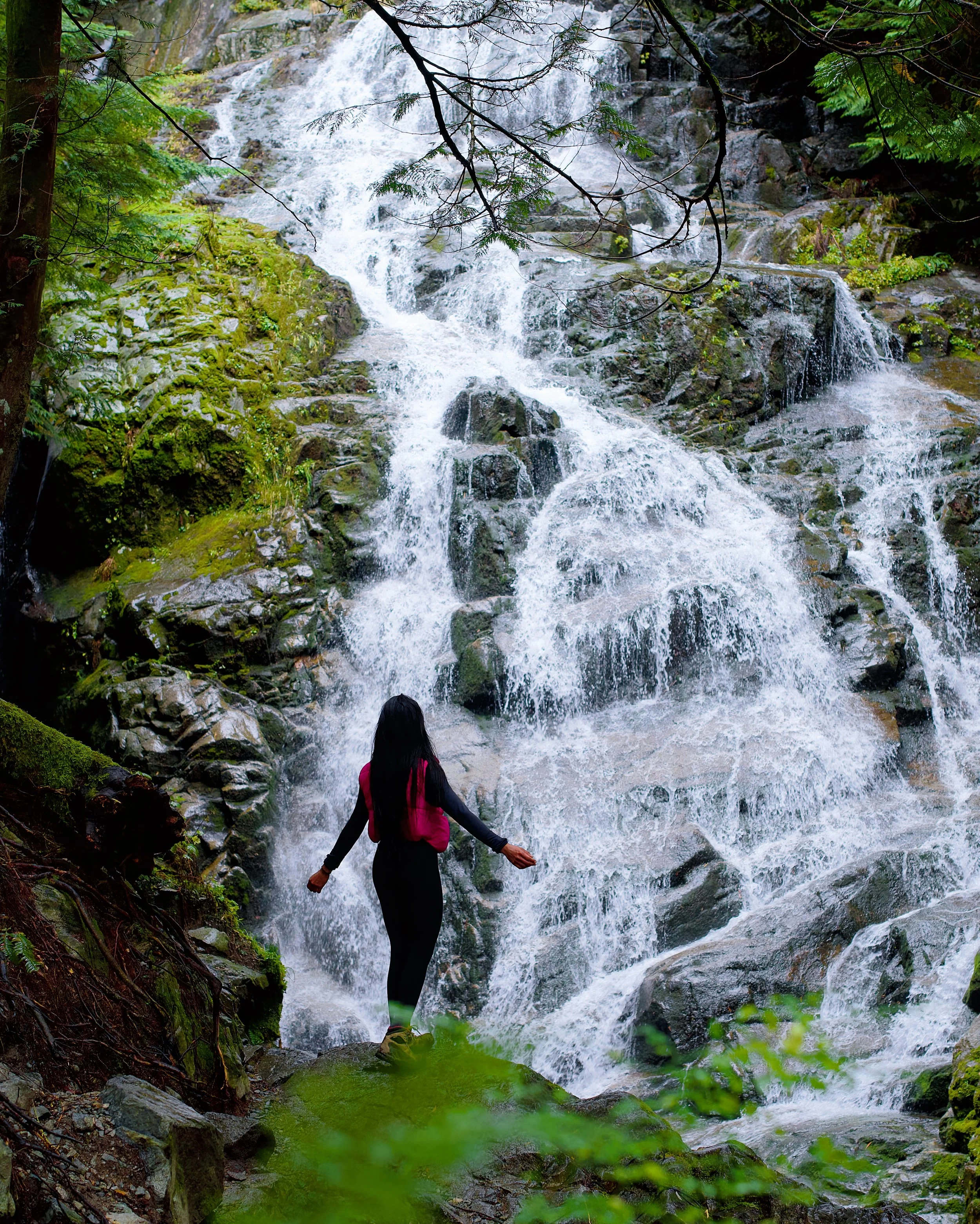 A woman stands in front of a waterfall in the forest, surrounded by green foliage and rocks.