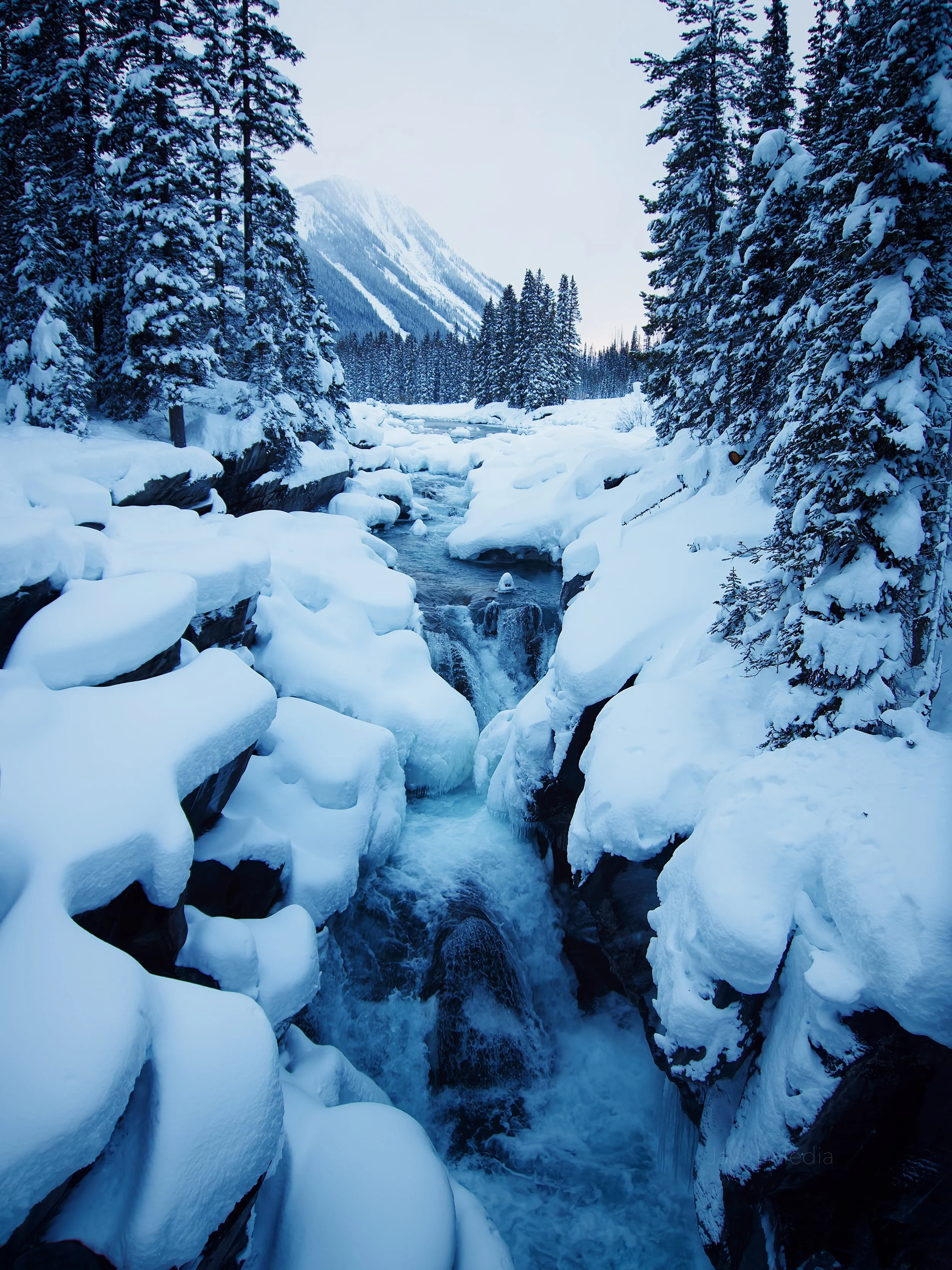 Snow-covered mountains and trees surround a rocky creek with flowing water in a winter landscape.