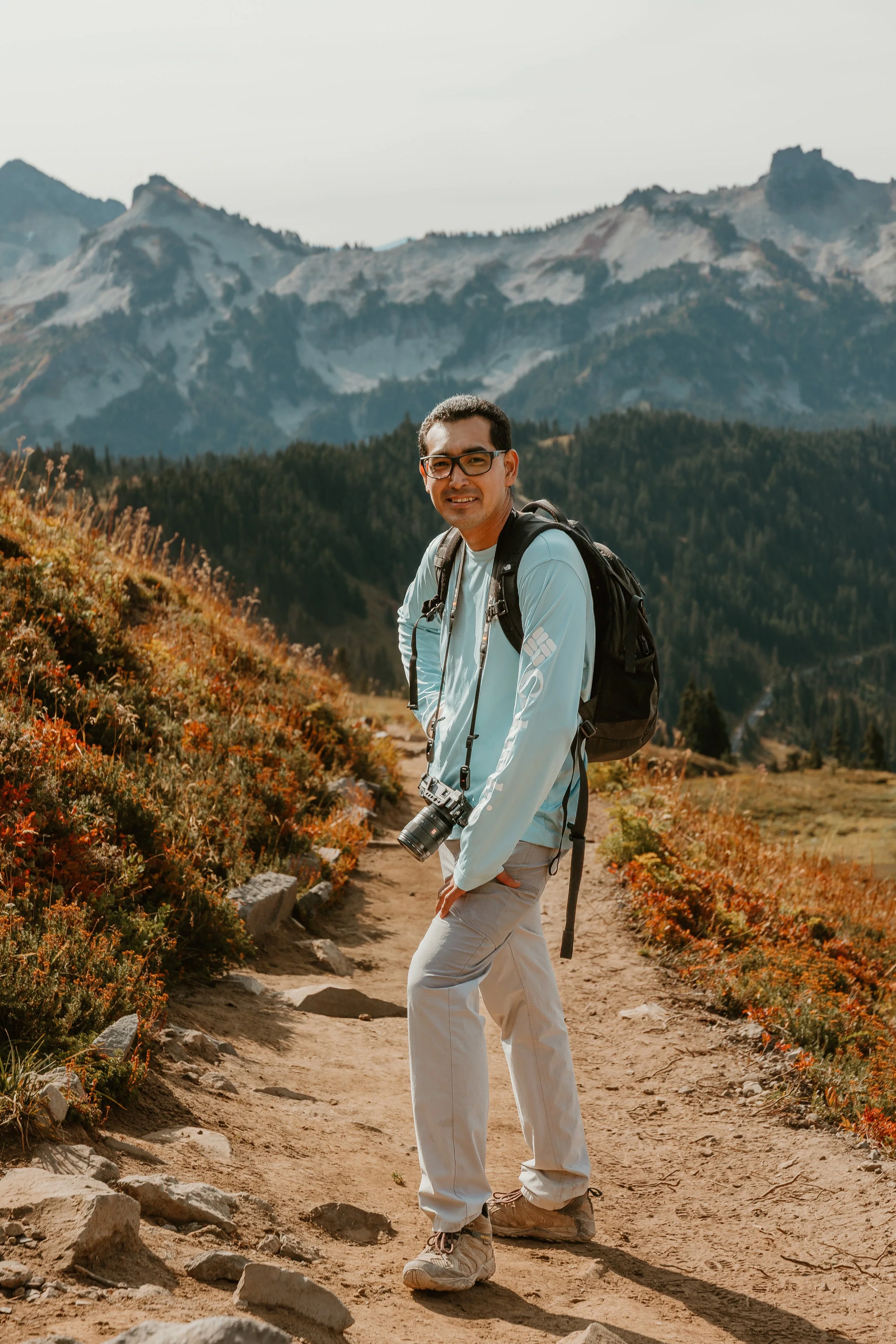 A man with glasses, wearing a light blue jacket and khaki pants, stands on a dirt trail in a mountainous landscape at sunset. He has a black backpack and a camera hanging around his neck, smiling at the camera.