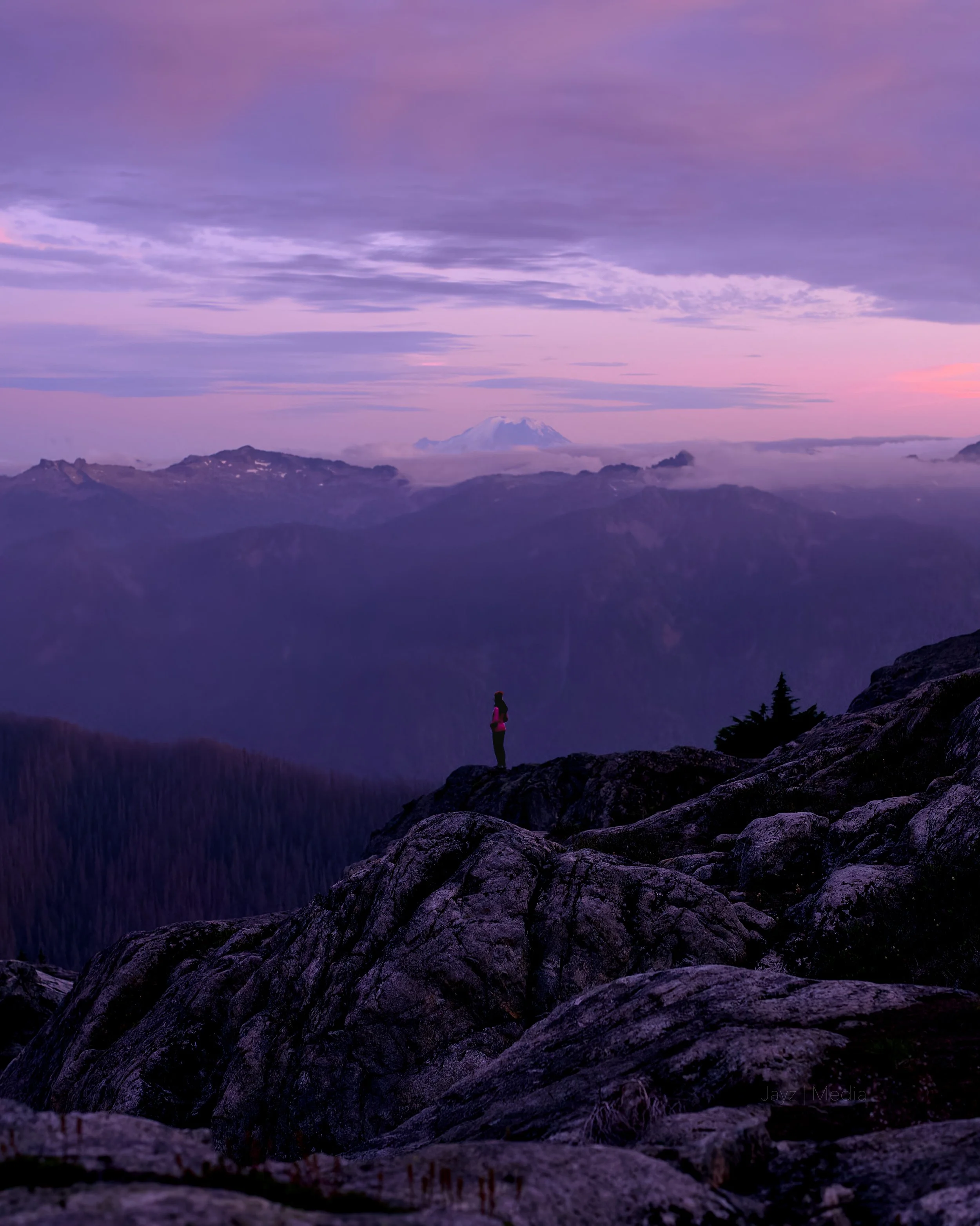 A person standing on rocky terrain overlooking a mountain range at dusk, with clouds and a snow-capped mountain in the distance.