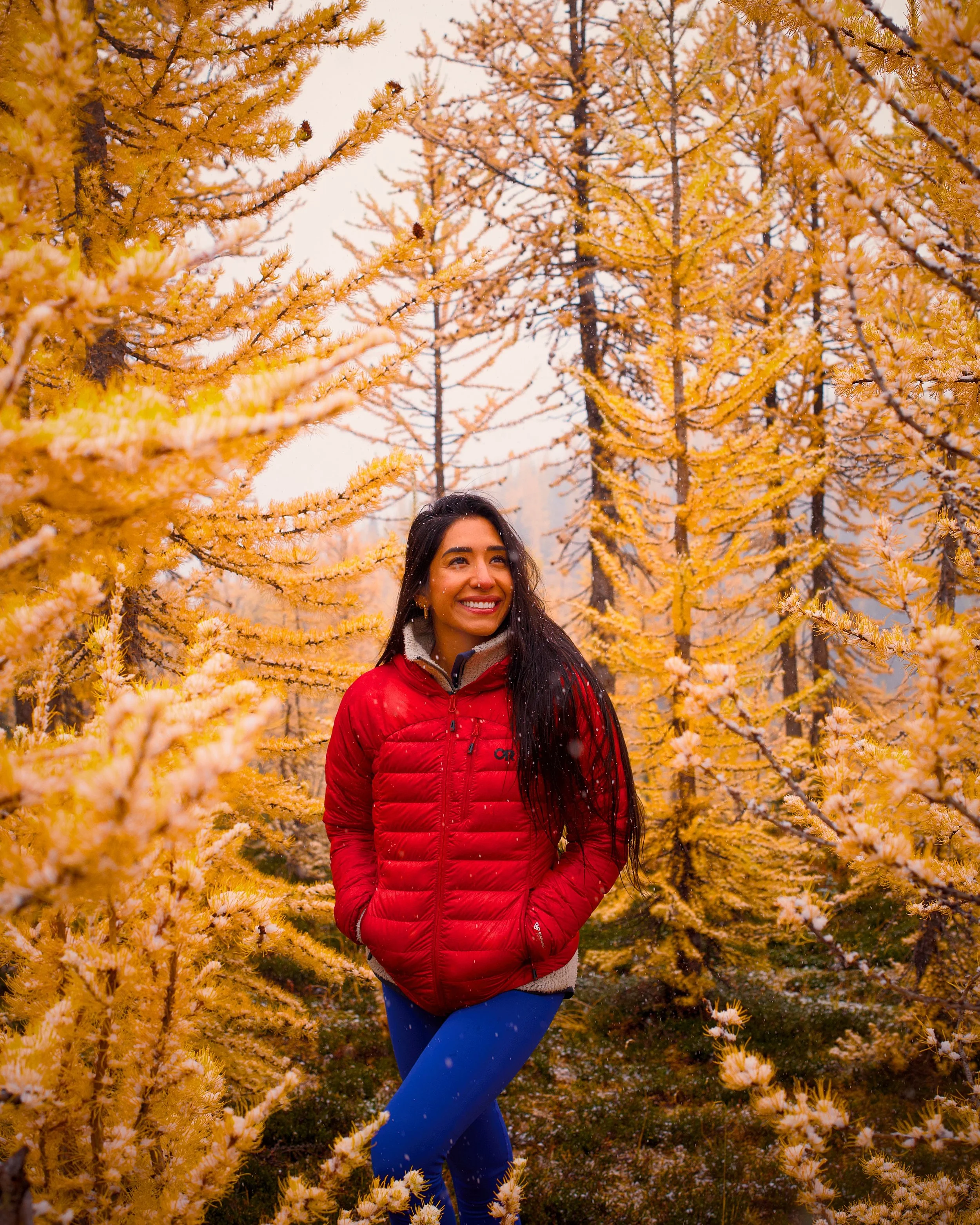 A smiling woman with long dark hair wearing a red puffer jacket and blue pants standing among yellow fall trees.