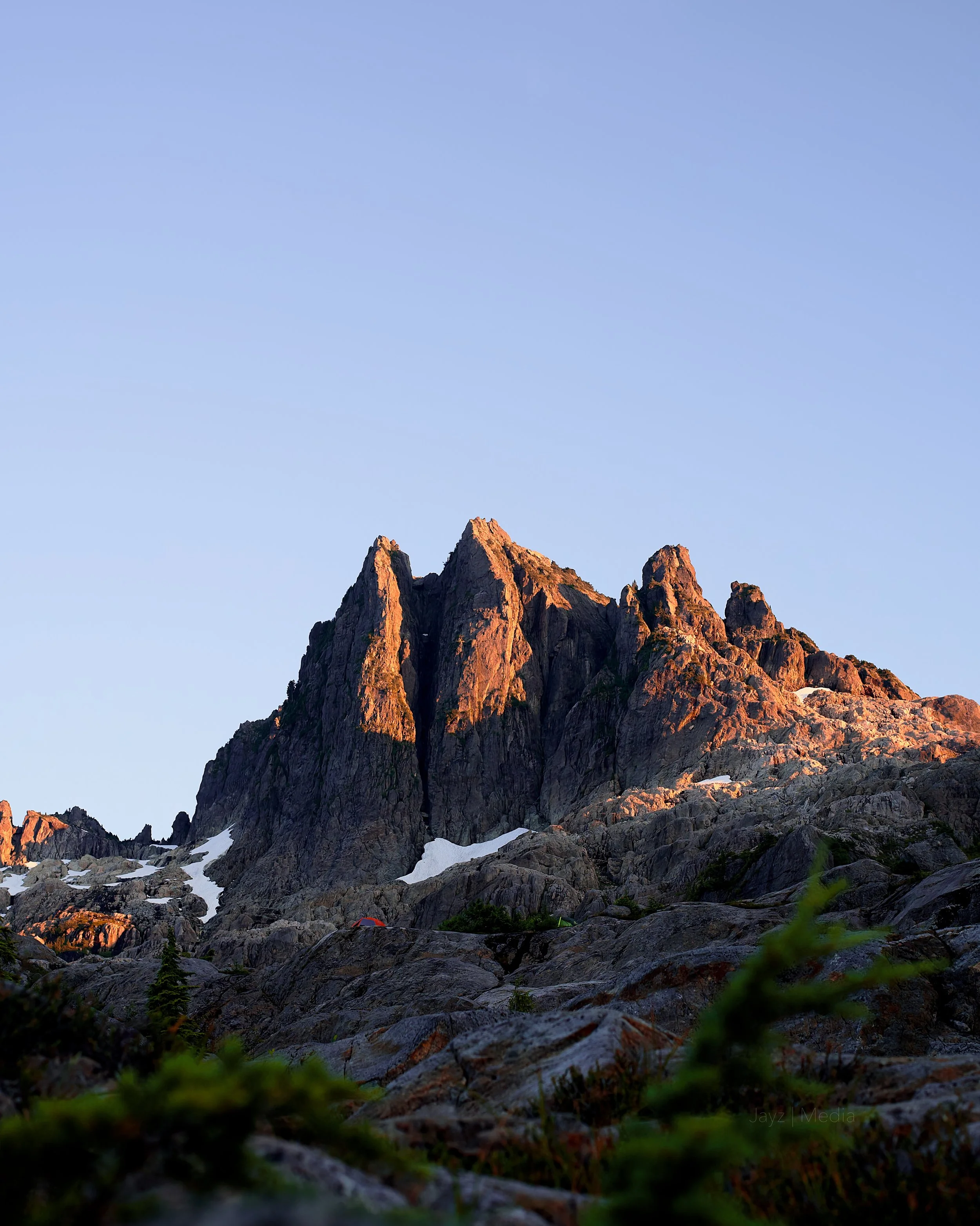 Sunlit mountain peaks with patches of snow, overlooking rocky terrain with sparse vegetation, under a clear blue sky.