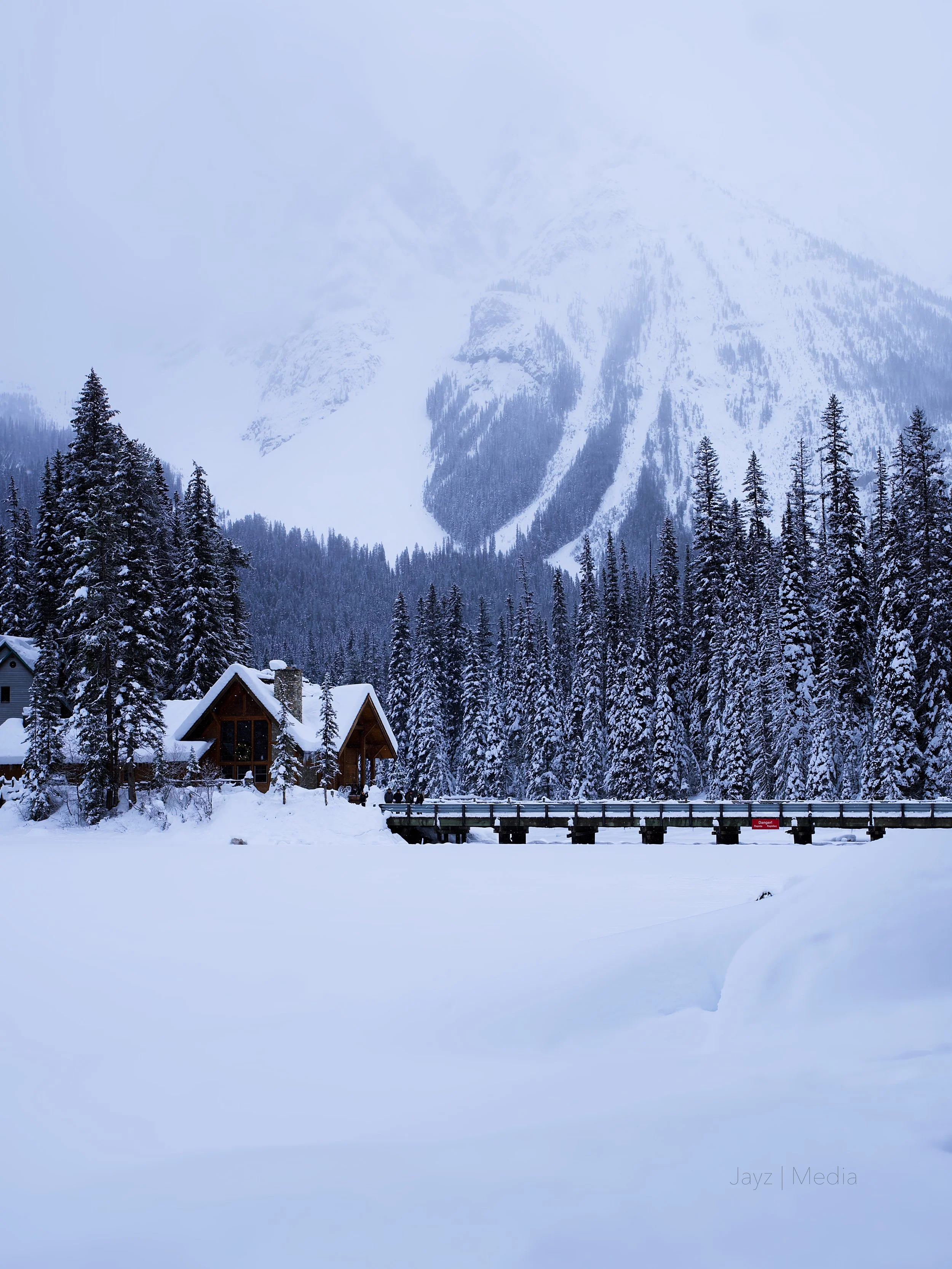 Snow-covered landscape with a wooden cabin, a bridge, tall pine trees, and snow-covered mountains in the background.