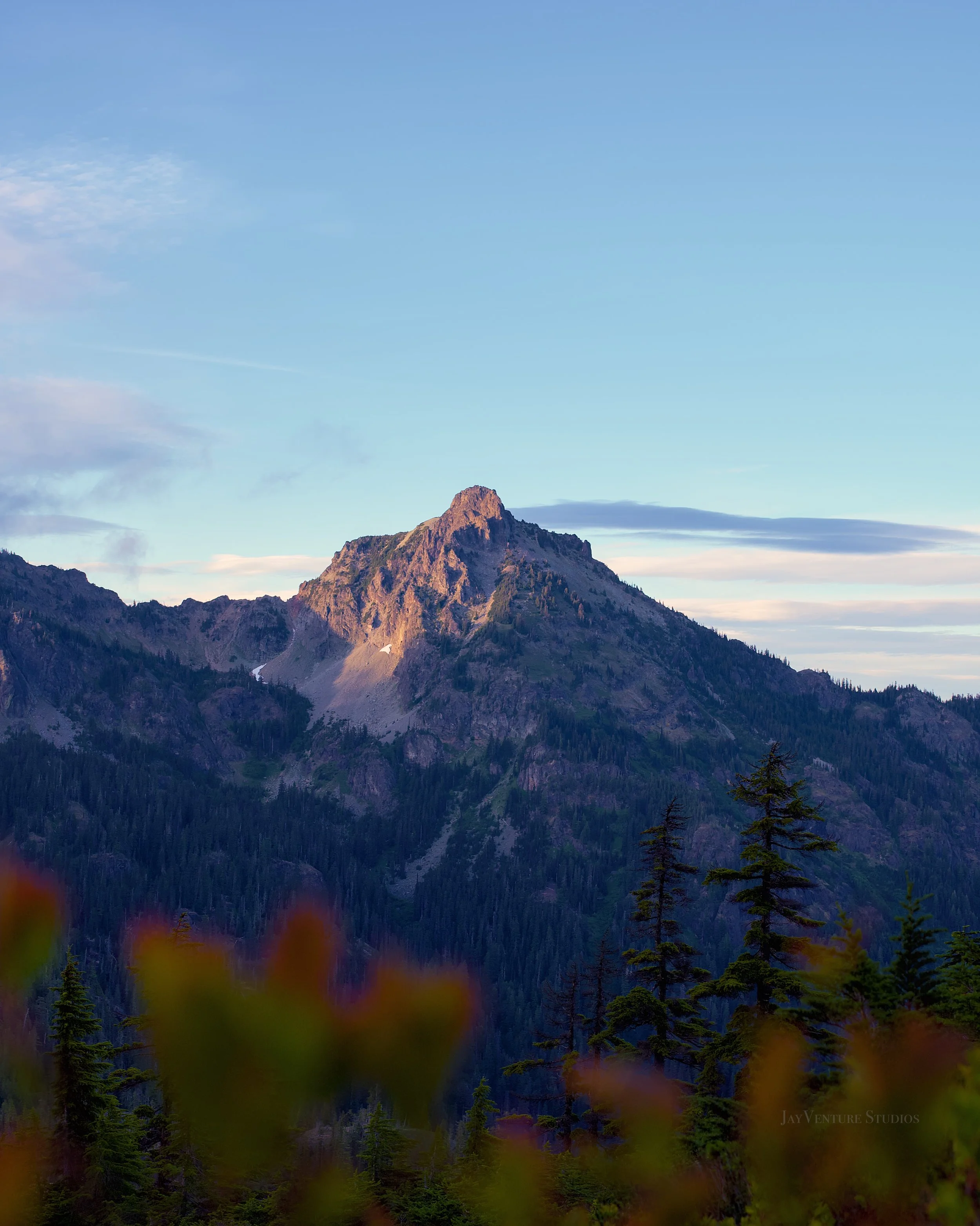 Scenic mountain landscape with a rugged mountain peak, trees in the foreground, and a clear blue sky with some clouds.