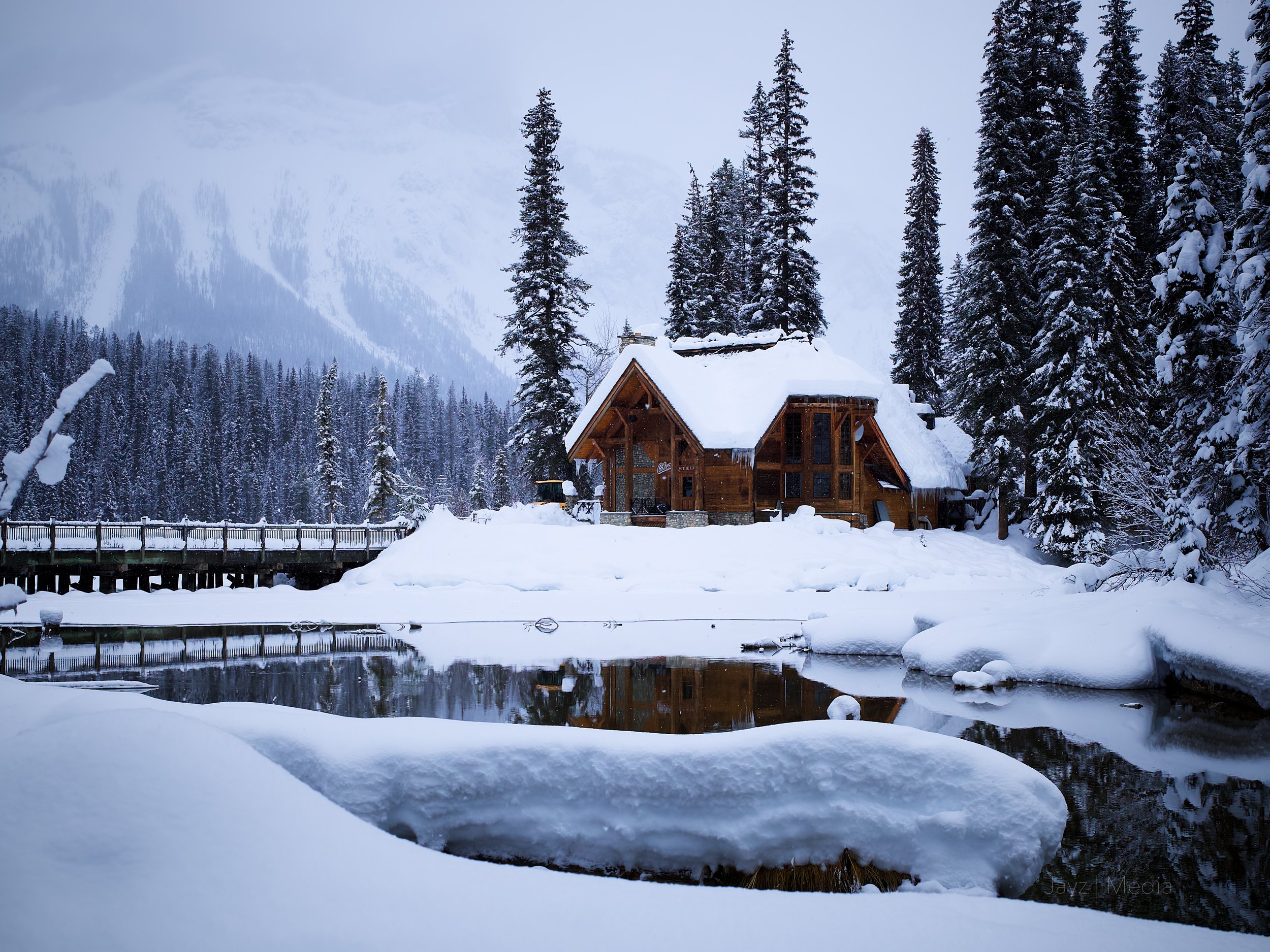 A snow-covered wooden cabin surrounded by tall pine trees and a frozen pond in a snowy forest landscape.