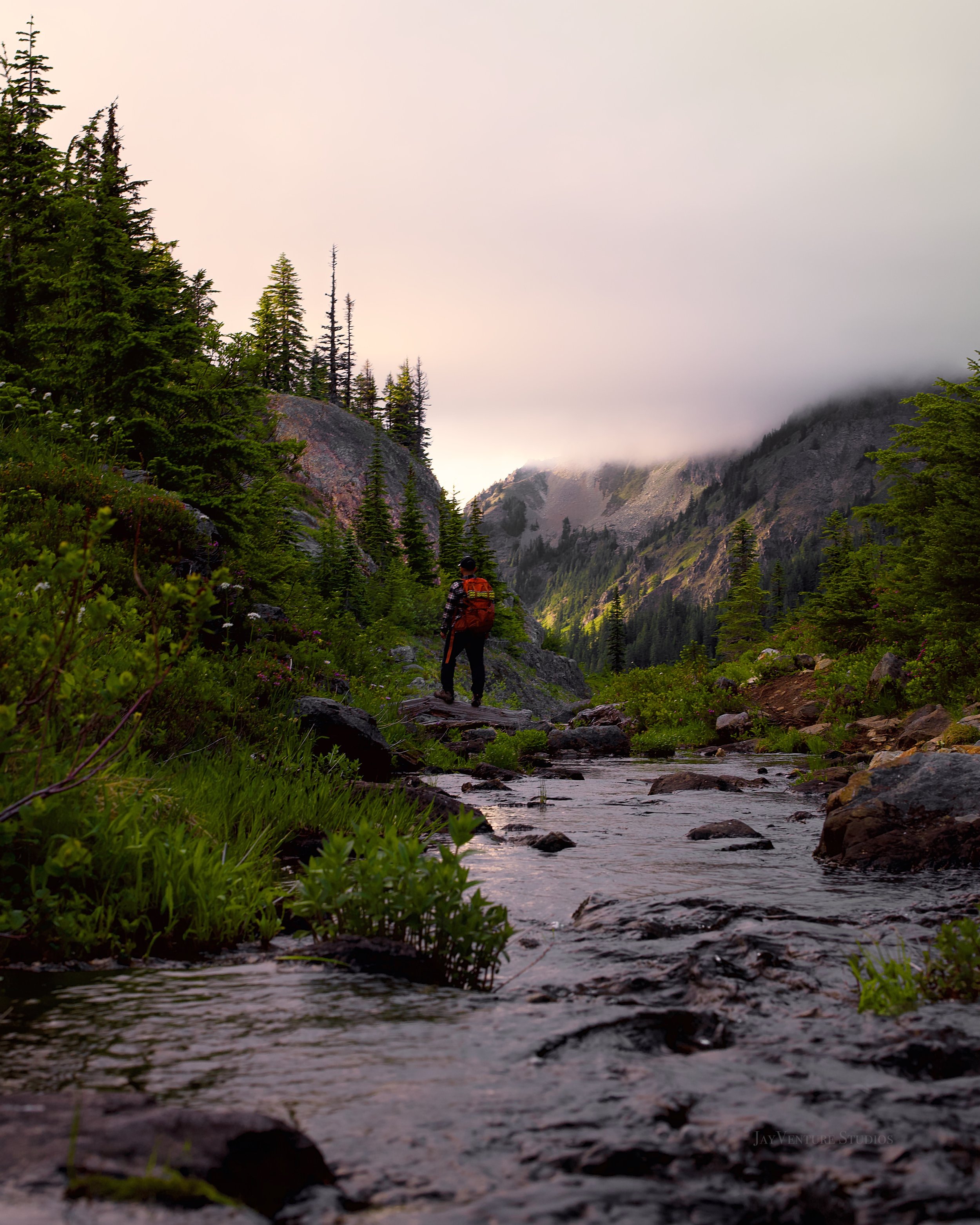 A hiker with an orange backpack walking along a rocky riverbank in a mountainous forest with tall trees and mist-covered peaks in the background.