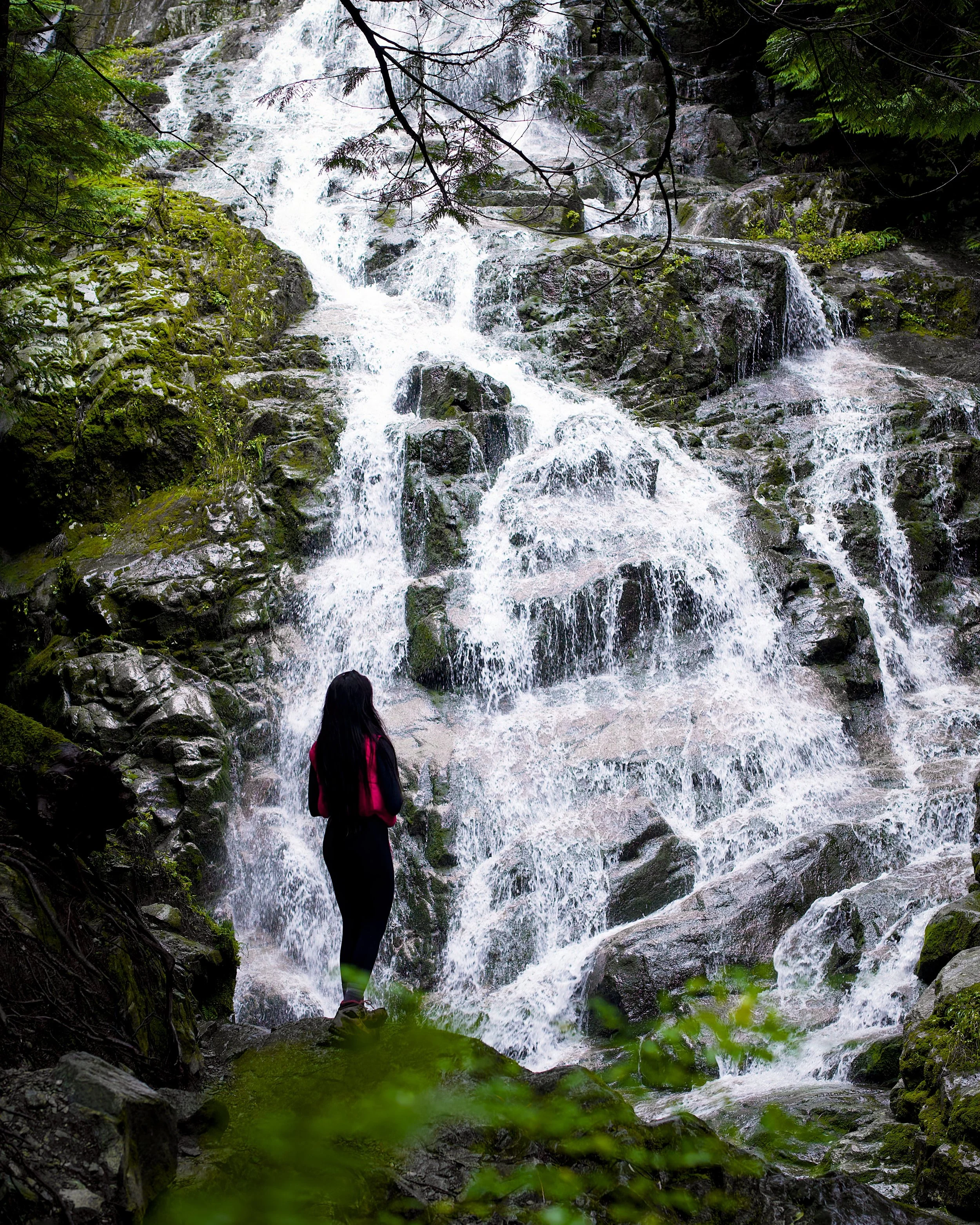 A person with long dark hair wearing a black outfit and red jacket standing in front of a rocky waterfall in a forest.