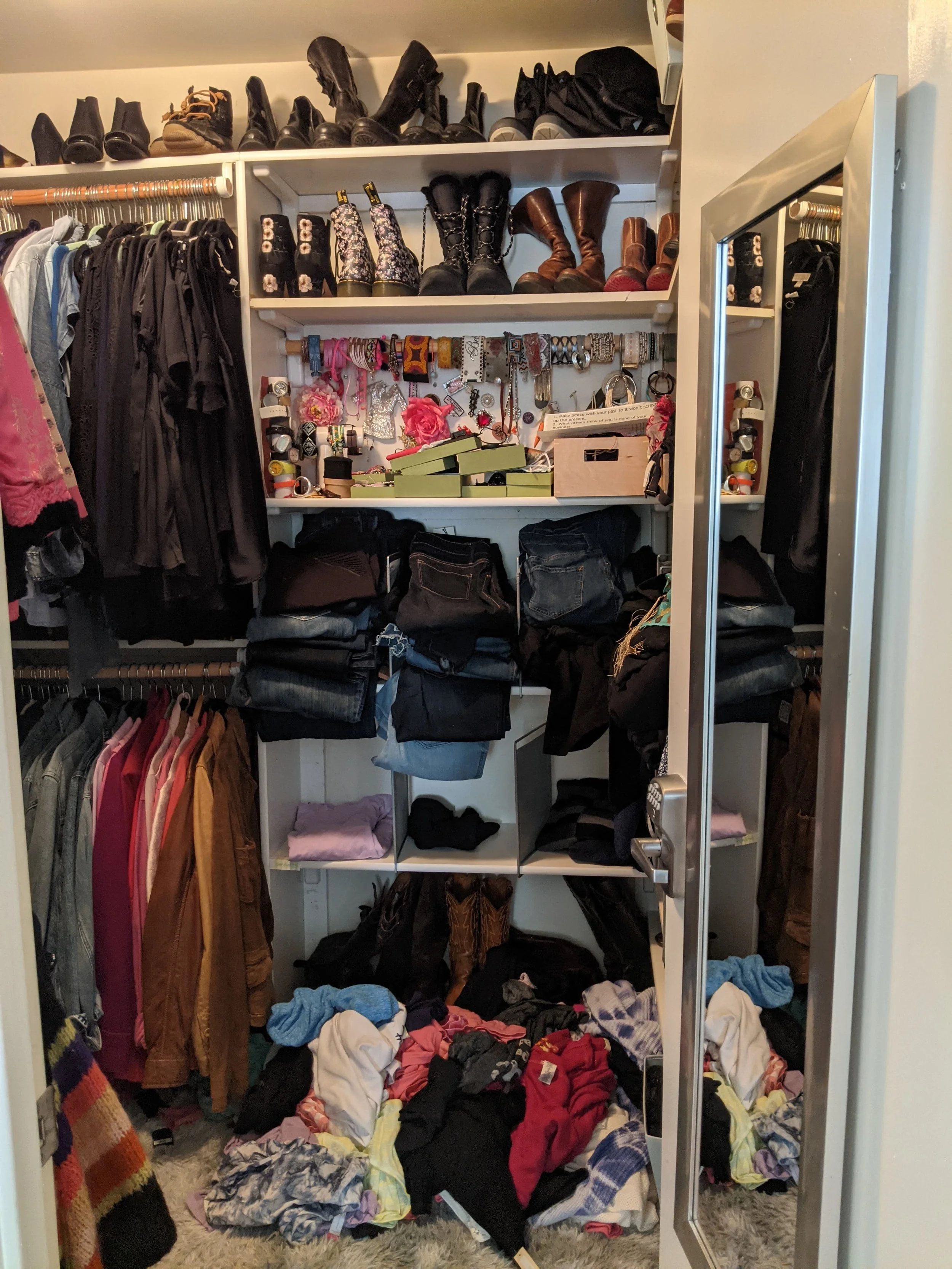 A cluttered closet with clothes hanging on a rod, shoes on top shelves, jewelry and accessories on a wall on the right, and clothes and shoes on the bottom. Clothes and items are scattered on the floor.