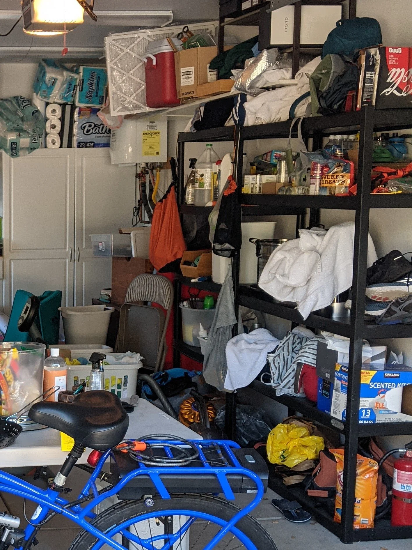 A cluttered garage with shelves filled with various household items, bags, boxes, and cleaning supplies, a blue bicycle in the foreground, and a white table with miscellaneous objects on it.