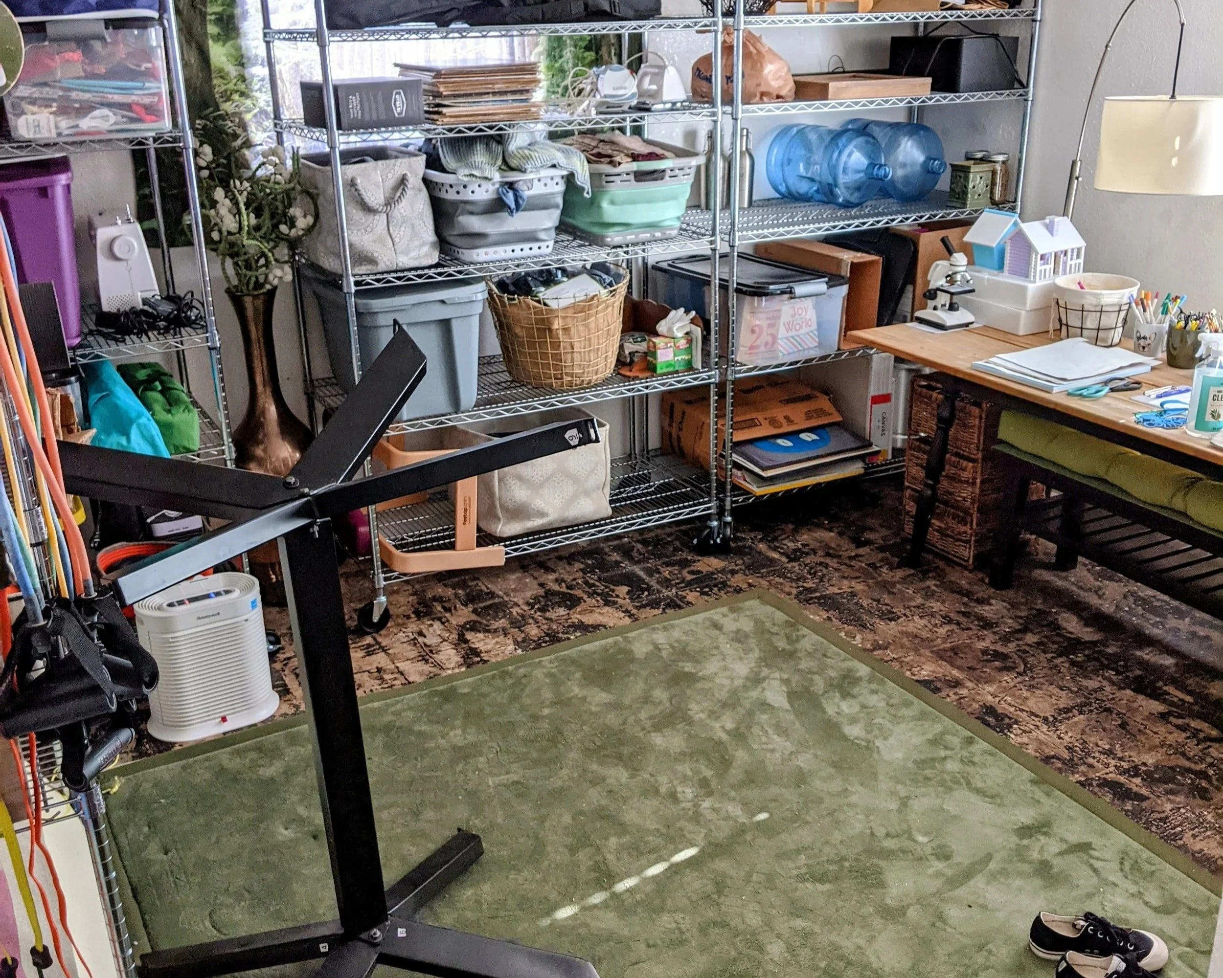 A cluttered garage with metal shelves filled with various household items, water bottles, baskets, and boxes. A work table with papers, pens, and a microscope. A small heater or dehumidifier in the foreground. Door and window with outside greenery visible.