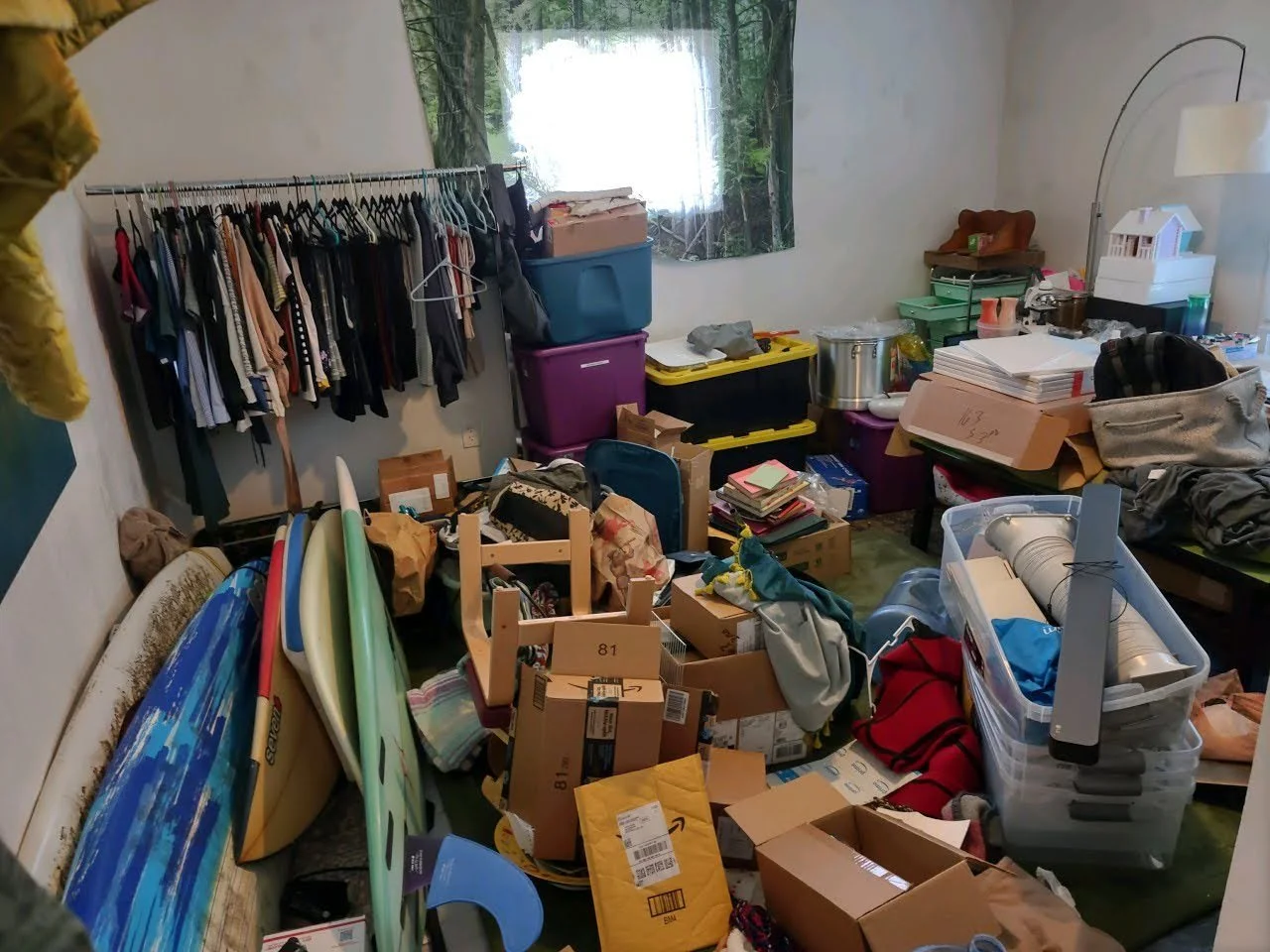 Room cluttered with clothes, cardboard boxes, and various household items including surfboards, papers, and storage containers.