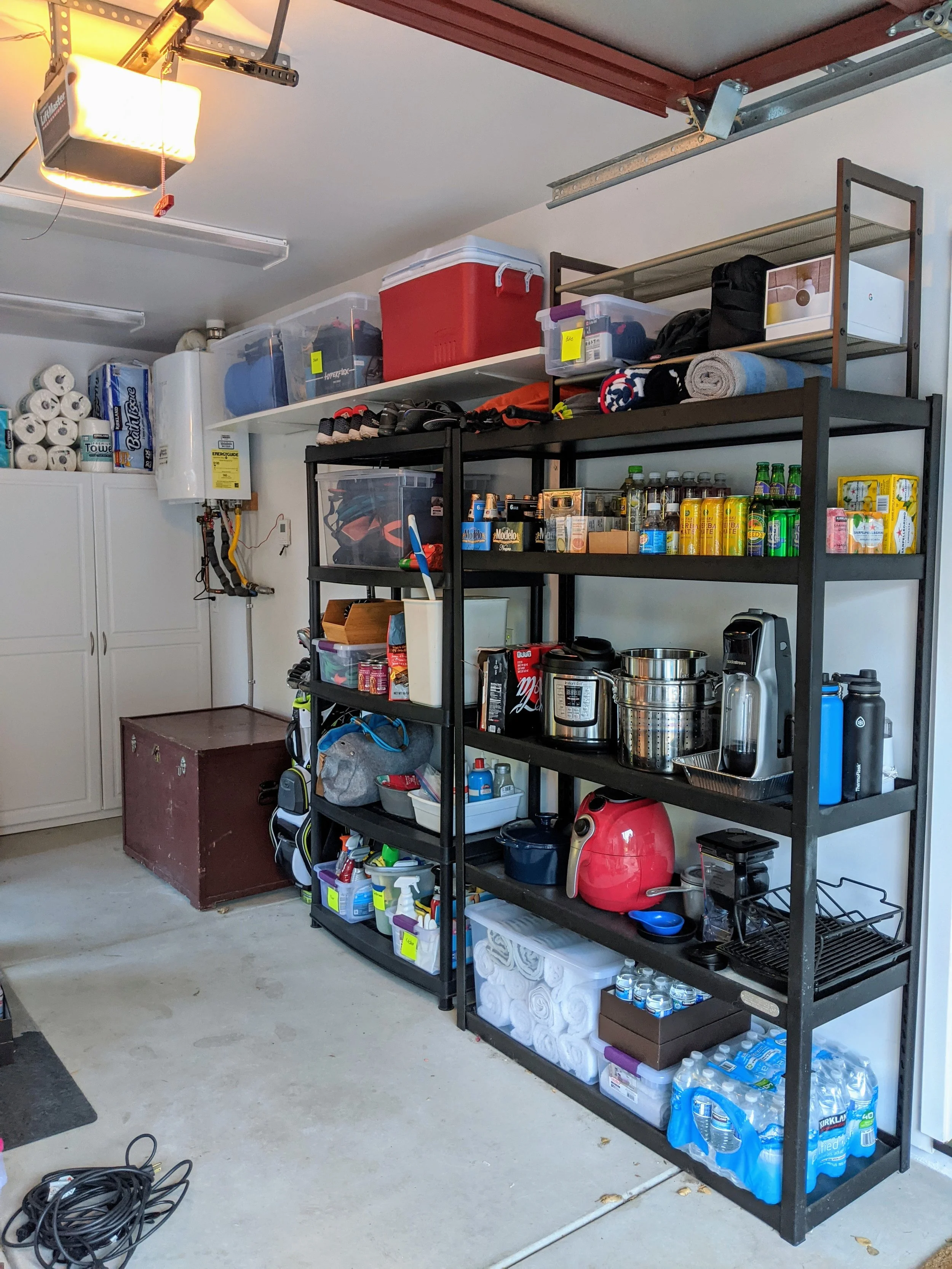 A garage storage area with a metal shelving unit holding various household items, beverages, and cleaning supplies, with a garage door opener on the ceiling and a large chest at the bottom of the shelving unit.