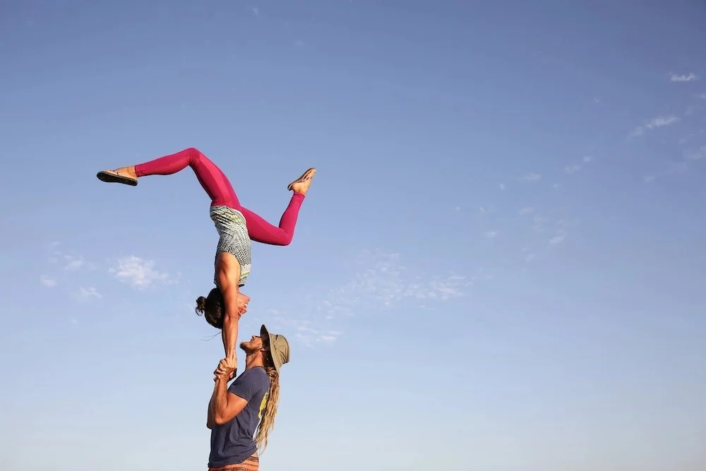 Two people set against a blue sky are practicing yoga. The man stands in profile with his hands near his shoulders. A woman is in a handstand facing him, but upside down.
