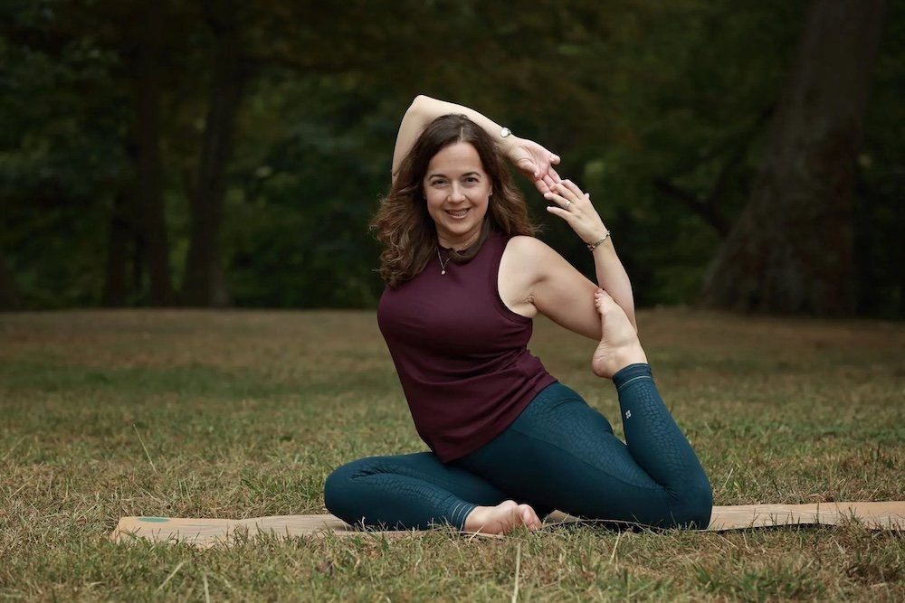 Woman practicing a seated yoga twist pose on a mat outdoors in a grassy park with trees in the background.