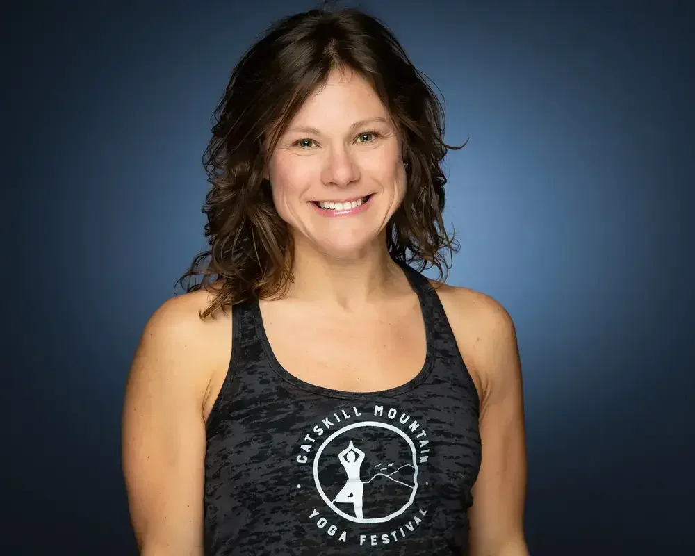 Smiling woman with shoulder-length curly brown hair, wearing a black athletic tank top with a logo that says 'Catskill Mountain Yoga Festival' against a dark blue background.