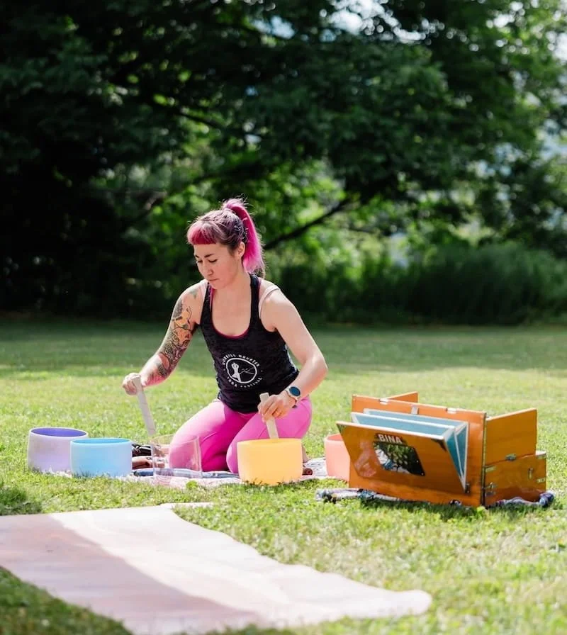 A tattooed woman with pink hair plays crystal singing bowls on a grass lawn outdoors, seated beside a harmonium and colorful bowls.
