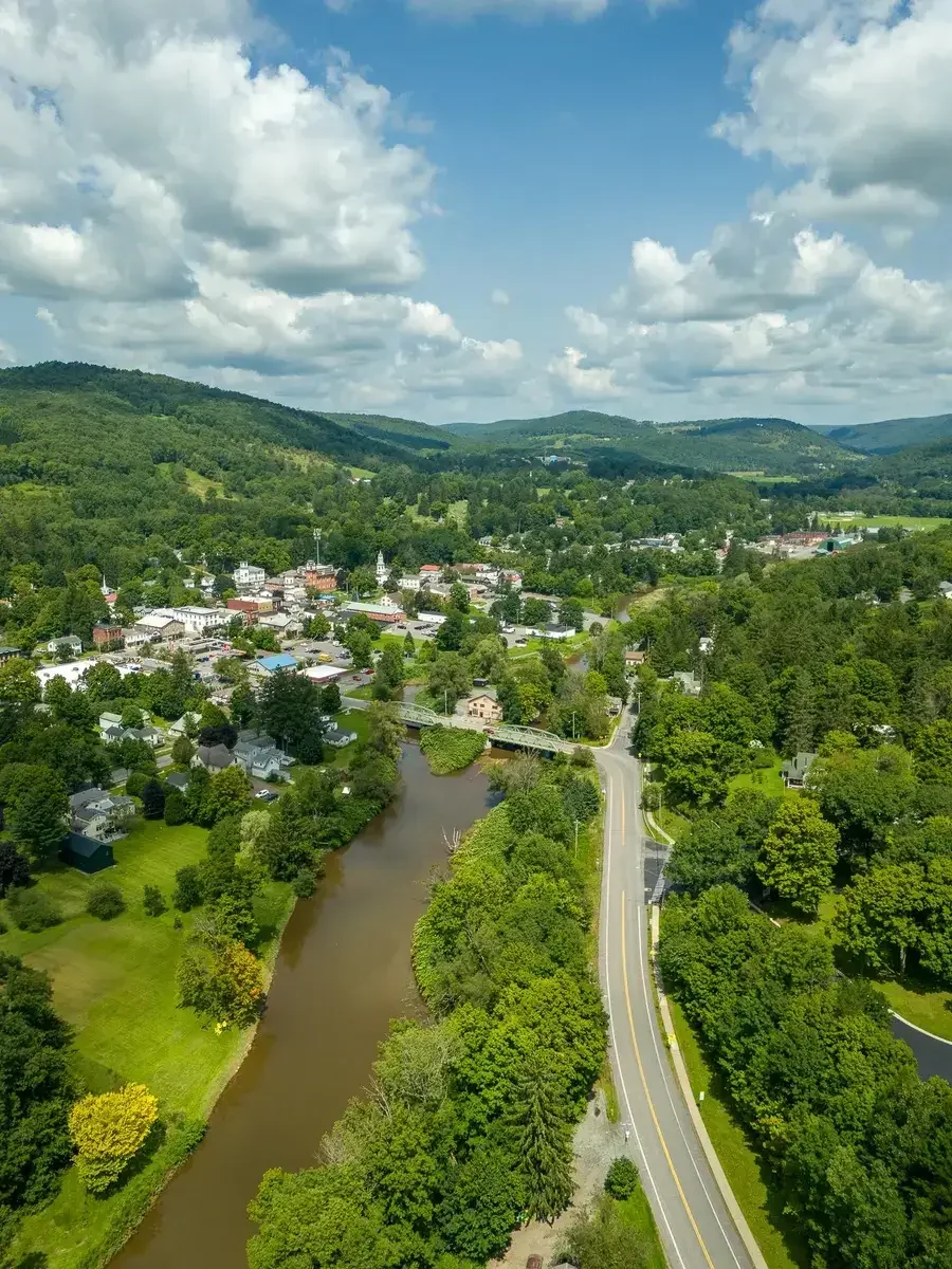 Aerial view of a small town with a river running through it, surrounded by green trees and rolling hills under a partly cloudy sky.