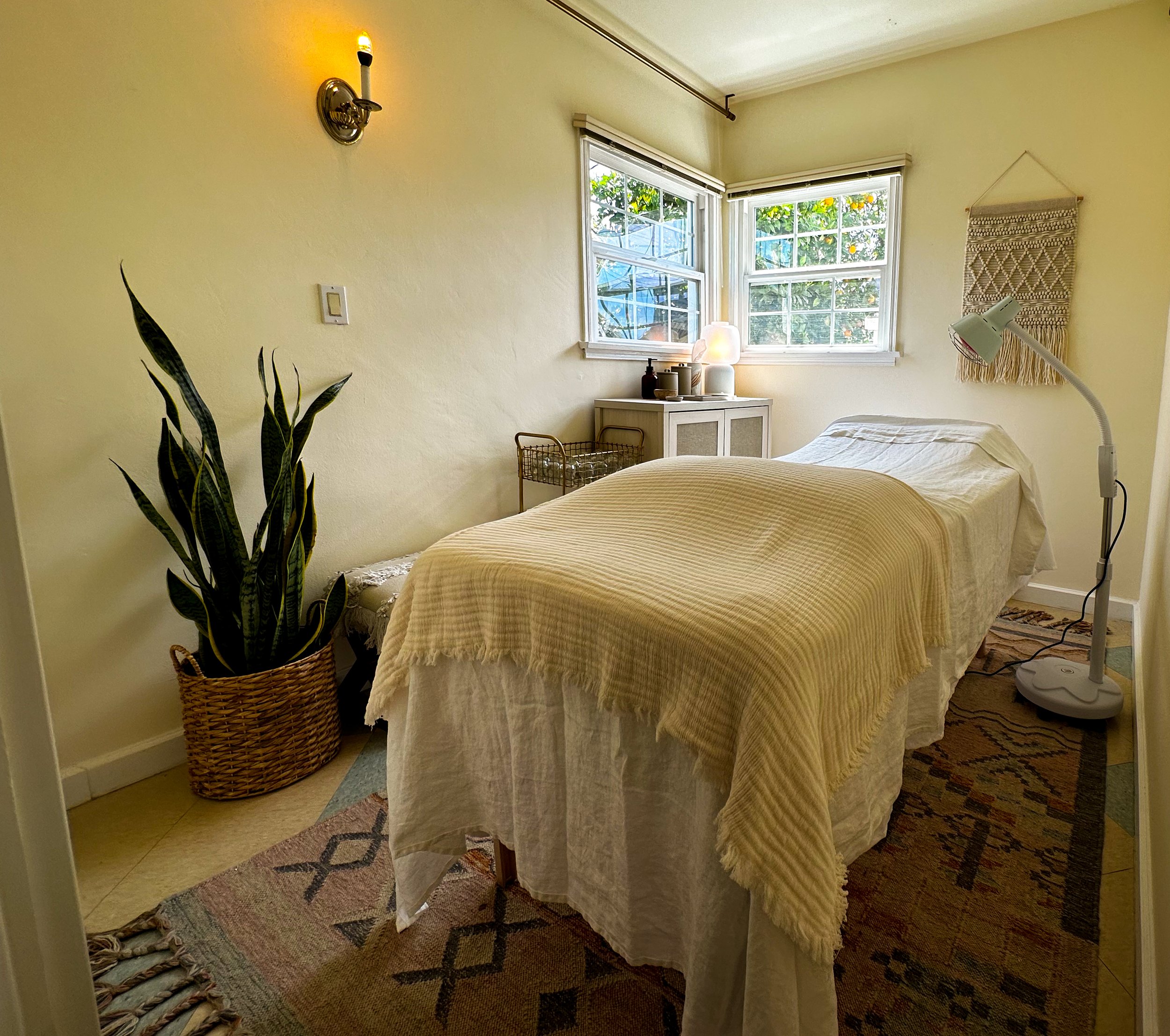 Peaceful treatment room equipped with massage table, infrared heat lamp, and view of orange trees in the backyard.