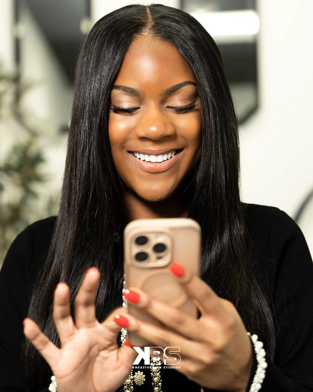 A smiling African American woman with long black hair looking at her phone in an indoor setting.