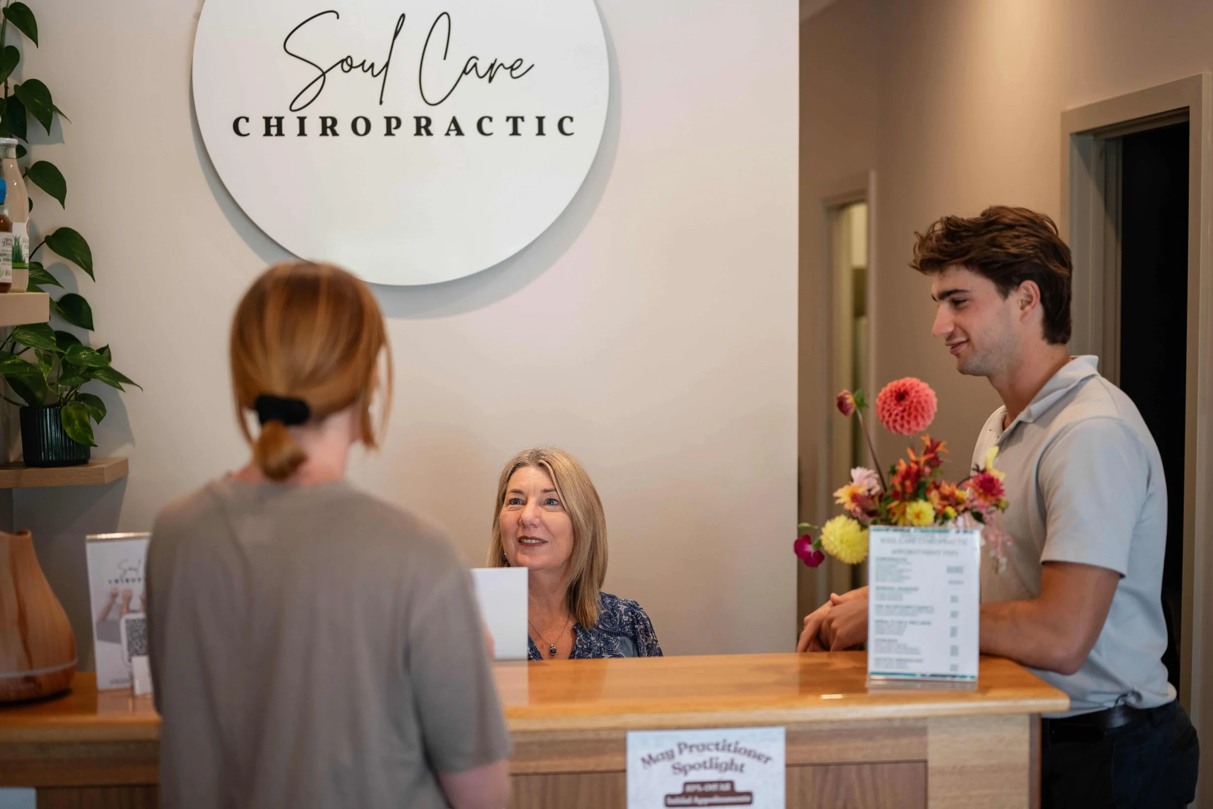 People check in at a chiropractor's office reception desk with a sign that reads 'Soul Care Chiropractic' in the background.