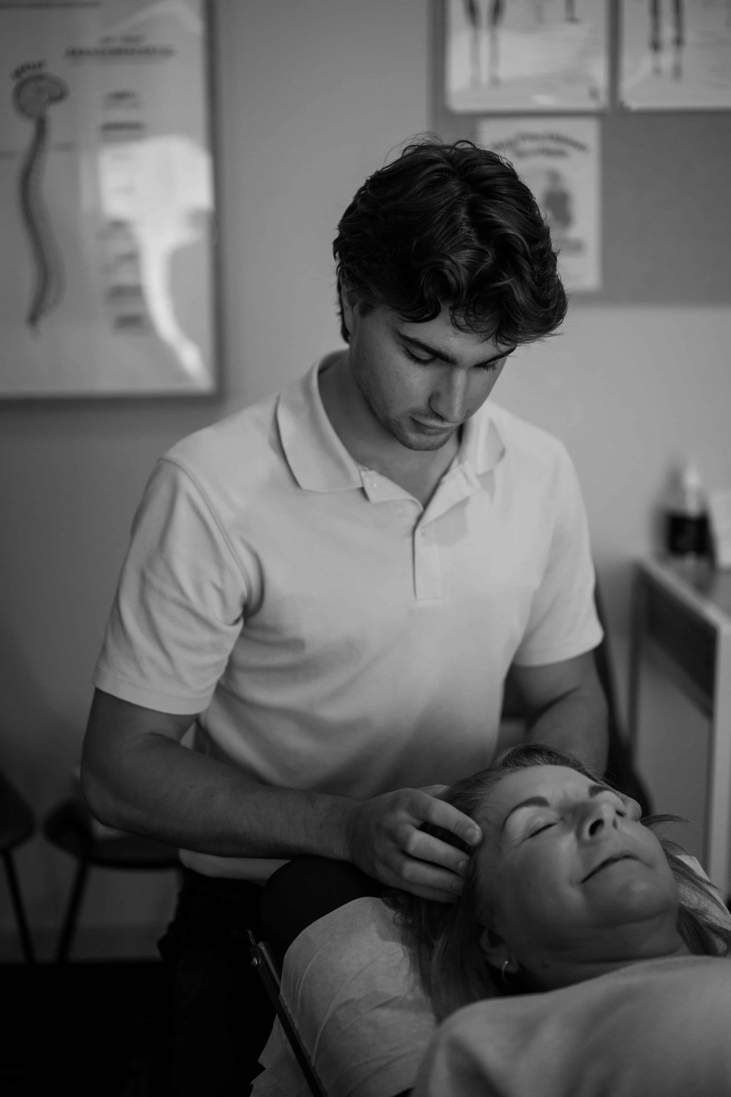 A man giving a massage to a woman lying on a massage table in a clinic. Both appear relaxed, and the setting includes medical posters on the wall.