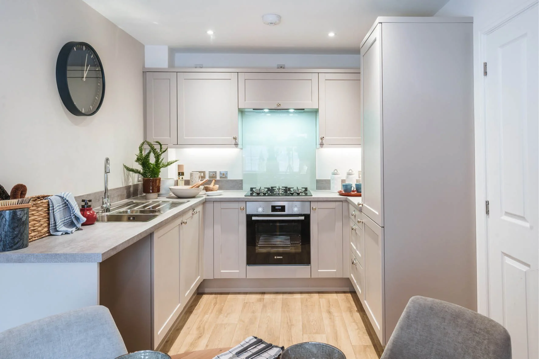 Modern kitchen with light gray cabinets, gas stove, stainless steel sink, wooden countertops, wall clock, and wooden flooring.