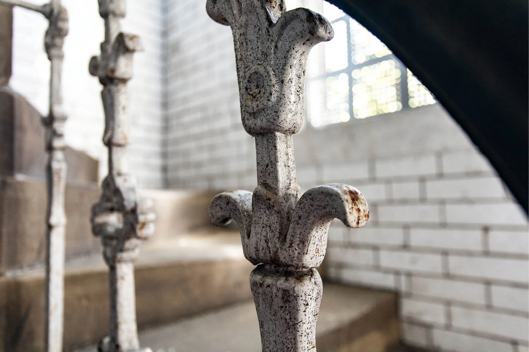 Close-up of decorative metal railing with rustic white paint and fleur-de-lis design, near brick wall and stairs.