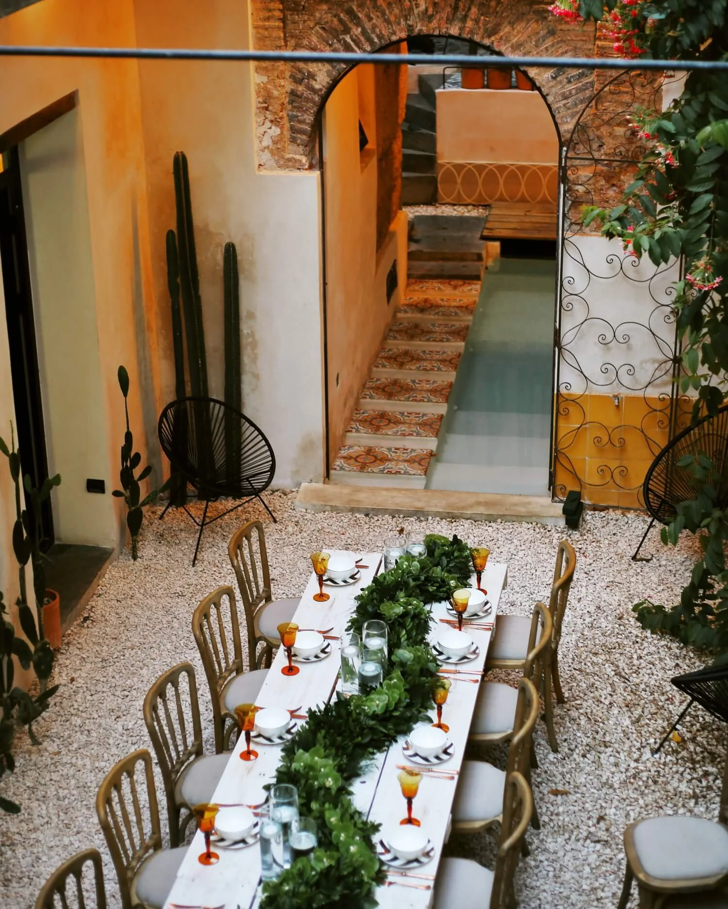 A decorated outdoor dining area with a long table set with plates, glasses, and a greenery centerpiece. Surrounding chairs are beige, and the area has a gravel ground. There are plants and a cactus on the shaded sides, and the background shows steps, brick arches, and colorful walls, creating a cozy, rustic atmosphere.