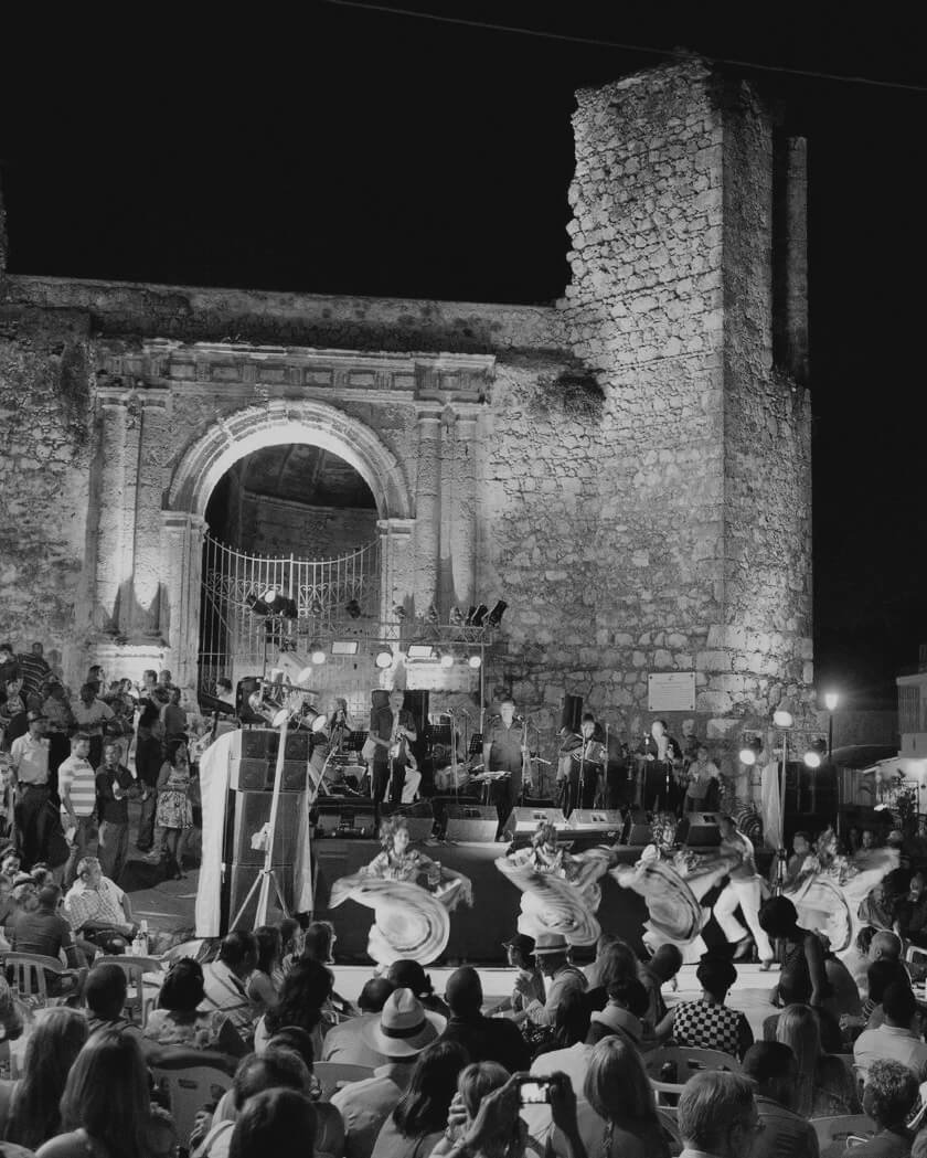 Nighttime outdoor concert in front of an ancient stone structure with an arch and tower, featuring musicians on stage, dancers performing in front, and a large audience watching.