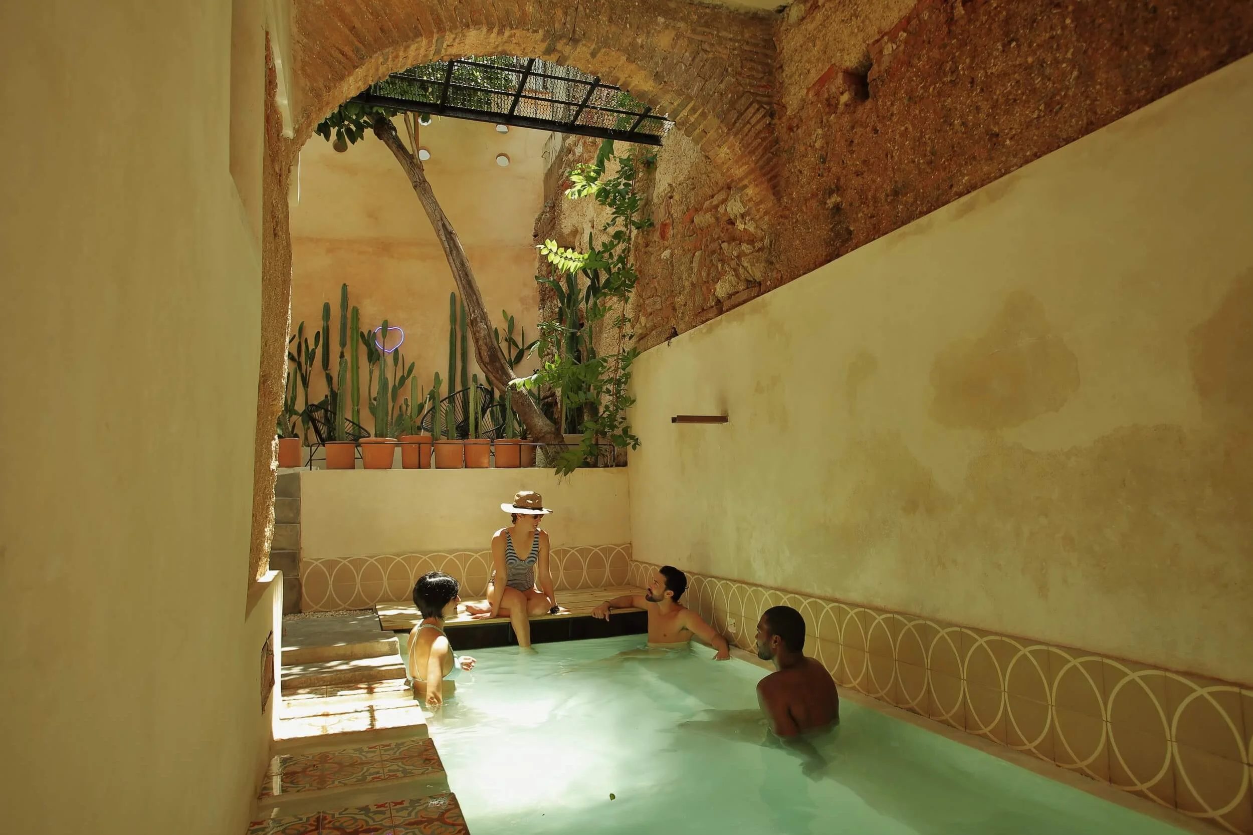 Group of friends relaxing in a small indoor pool under a historic stone arch with cacti in Zona Colonial Santo Domingo.